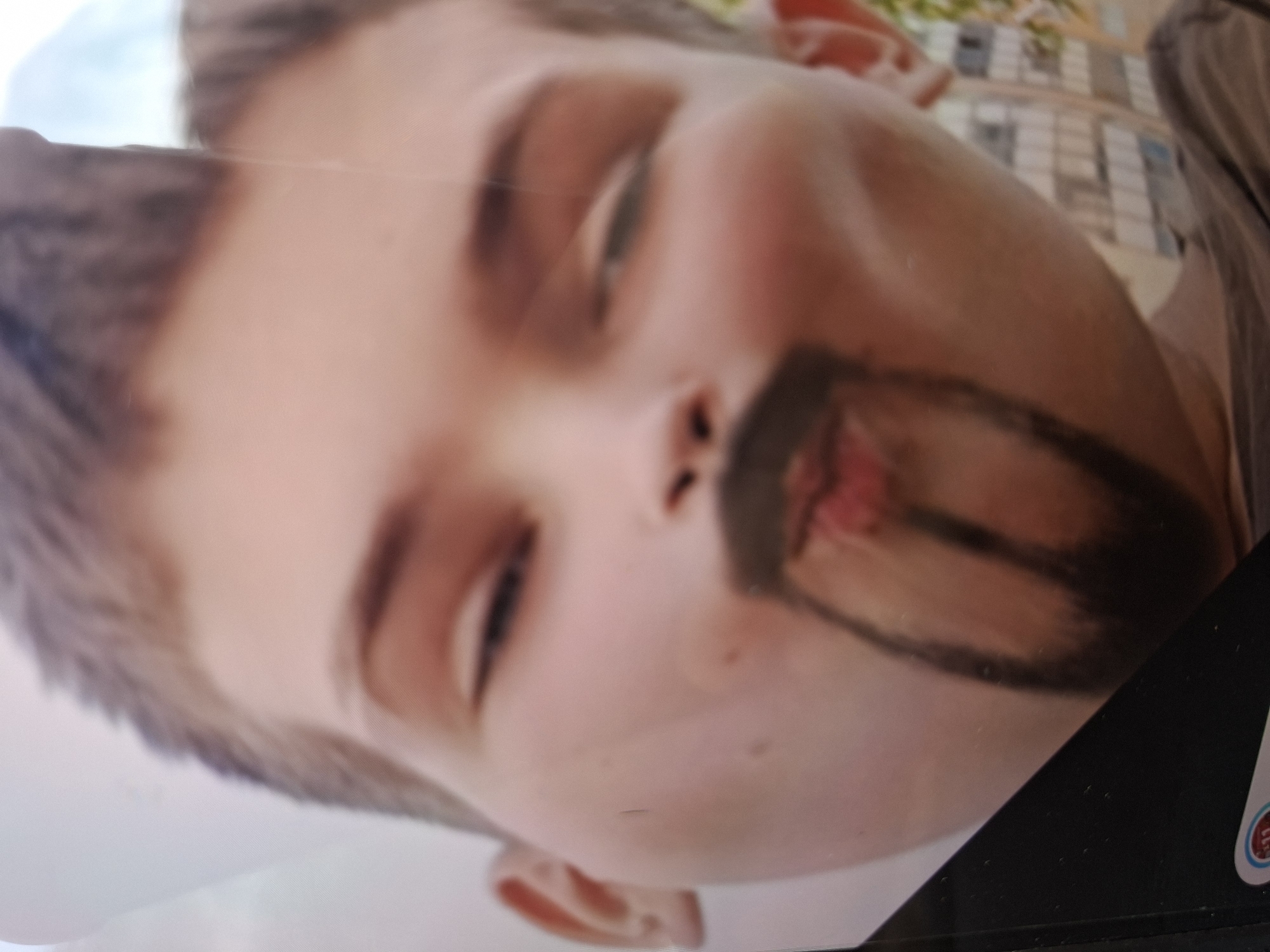 An outdoor photograph captures a close-up of a large advertisement or poster featuring a child's face, likely a boy due to short hair, tilted at an angle. The child has fair skin and closed eyes, conveying a serene or sleeping expression. Significant vandalism is present on the poster, with dark marker drawings creating a caricature of a goatee and an exaggerated, elongated tongue extending from the child's mouth down the chin. The "tongue" is a dark, somewhat rectangular shape with a reddish center.
The poster is displayed in an urban outdoor setting, with blurry buildings visible in the background, suggesting a street or public area in Saint Petersburg, Russia. The image is a photograph of a physical display, showing some reflections and the texture of the printed material. A small, partially visible circular sticker with a red and white design and the text "12+" is discernable on the dark frame surrounding the poster in the bottom right corner. The overall impression is one of public property defacement.