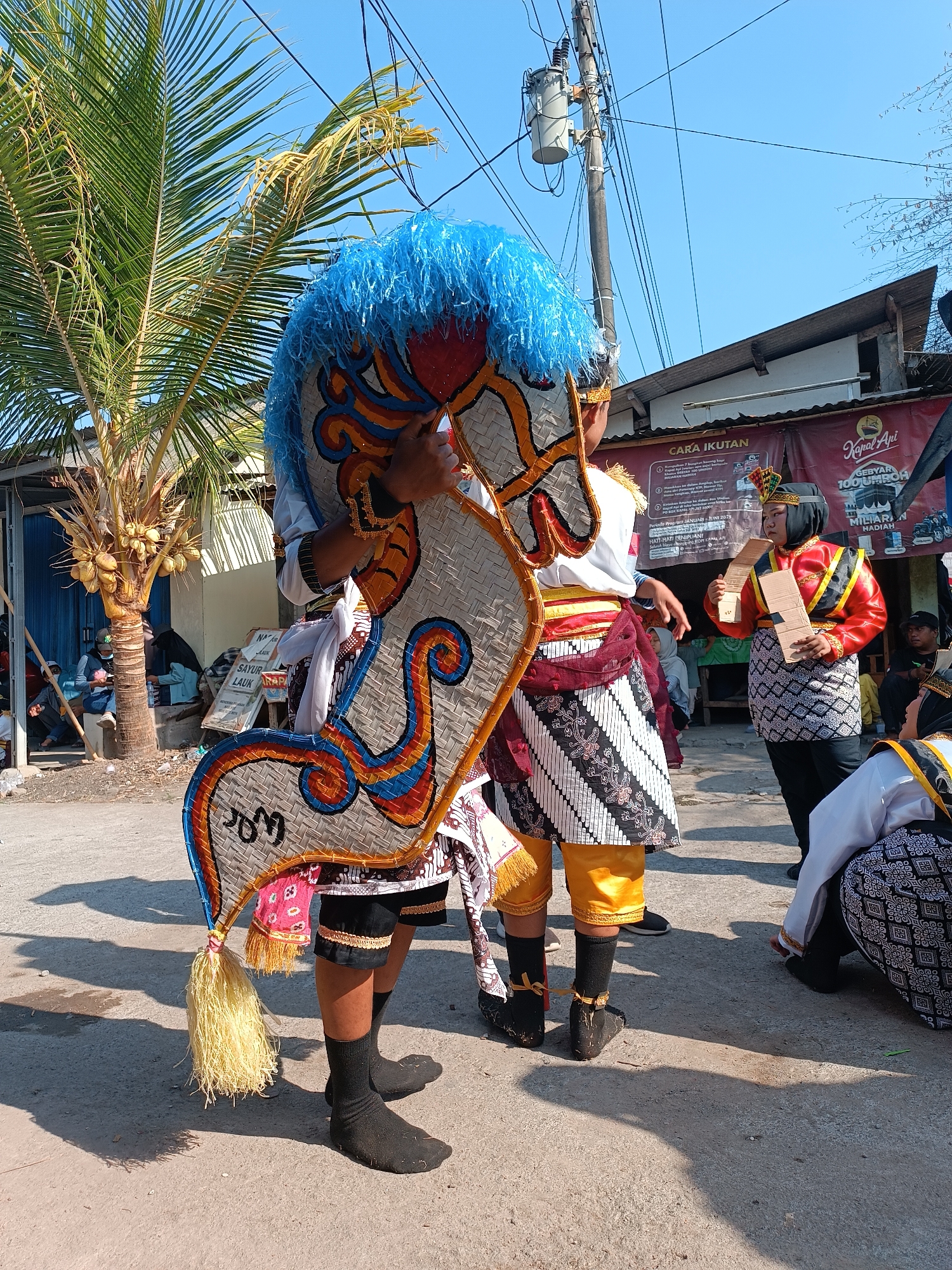 In Imogiri, Indonesia, under a clear blue sky, a cultural festival is underway. The foreground features two individuals participating in a traditional performance. One person, dressed in a white shirt with red and gold accents and a maroon sarong patterned with white batik, holds a large, ornate horse puppet. The puppet has a blue, feathery mane and a woven body decorated with colorful patterns. The other individual, wearing a red jacket with gold trim, a black headscarf, and a black and white patterned sarong, holds a piece of folded brown paper. Behind them, a palm tree laden with coconuts stands to the left, casting a shadow on the dusty ground. Further in the background, buildings with corrugated metal roofs line the street, and several spectators can be seen observing the festivities, some seated in the shade of an open-fronted building. Banners advertising events and products are also visible. The overall atmosphere suggests a lively community event taking place on a bright, sunny day.