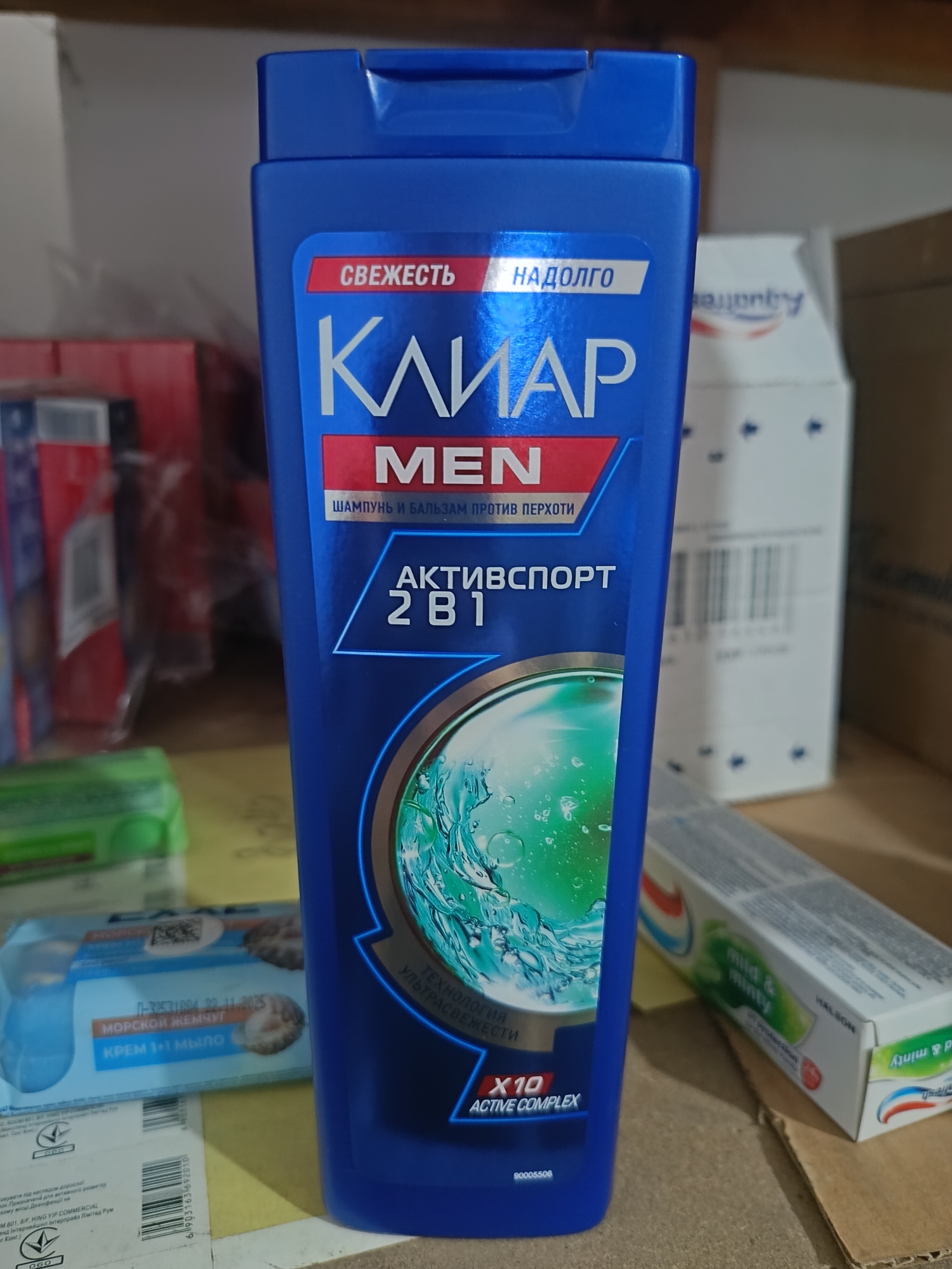 This image shows a close-up of a blue bottle of "Clear" brand shampoo for men, with Russian text on the label. The bottle is positioned on a surface, possibly a shelf or table, with other items visible in the background, including a box of toothpaste and what appear to be other product boxes or packaging. The lighting is direct, highlighting the glossy surface of the shampoo bottle. There is no visible indication of the city of Berdiansk, Ukraine, or any environmental details that would place the scene in a specific urban location. The focus is entirely on the product and its immediate surroundings.