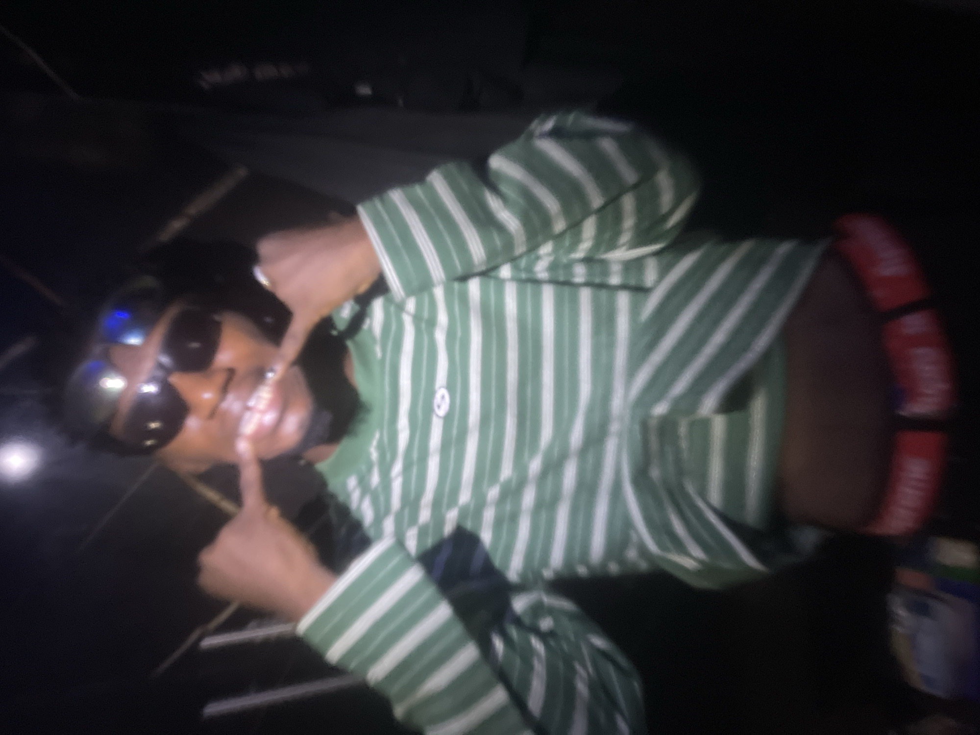 A person is pointing at their teeth in a blurred image. The person is wearing sunglasses and a green and white striped shirt. It is not possible to determine the location in Lokoja, Nigeria, based on the image provided.