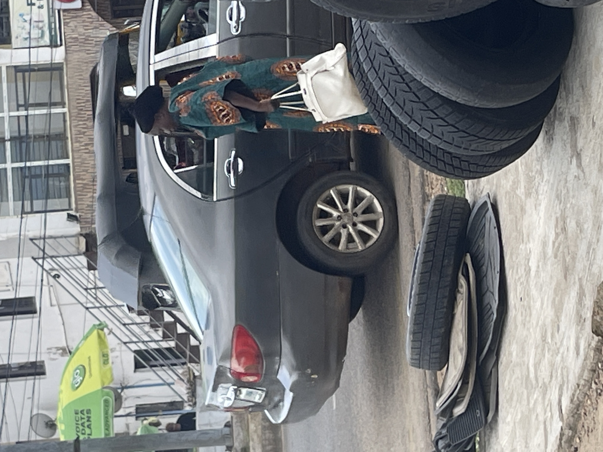 In an outdoor urban setting, a dark-colored sedan is parked on a street next to a sidewalk. A woman wearing a black headscarf and a vibrantly patterned traditional outfit stands to the right of the car, holding a large white handbag. In the foreground, several car tires and floor mats are stacked on the sidewalk. In the background, a building with multiple windows and a set of stairs leading to an upper level is visible, along with a green and yellow umbrella advertising "Voice Data Plans." A three-wheeled auto-rickshaw is parked behind the sedan. The lighting suggests it is daytime.