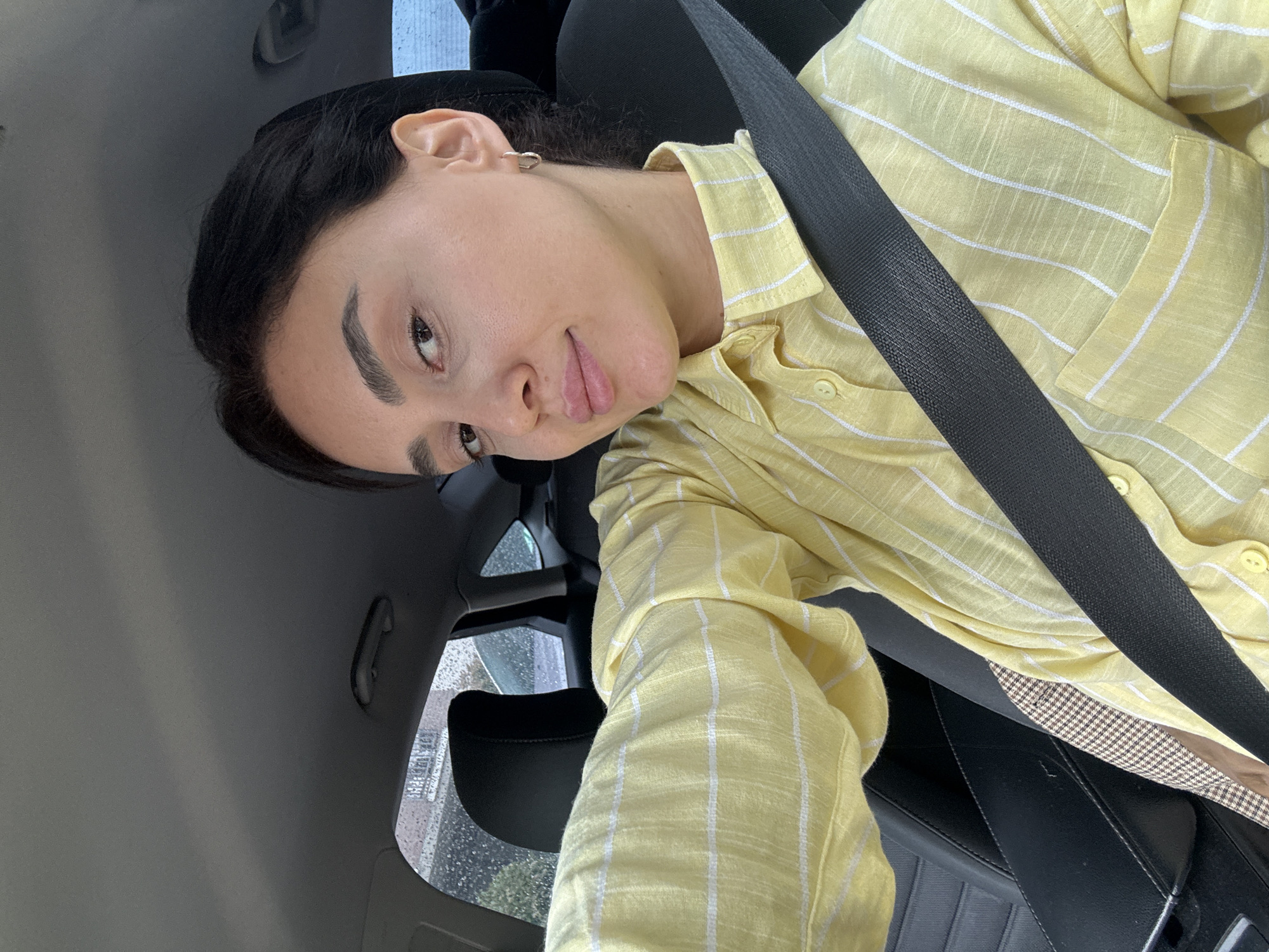 A woman is seated in the driver's seat of a car, wearing a yellow striped shirt and a seatbelt. The car appears to be moving, given the rain-streaked window behind her. The woman is looking directly at the camera with a slight smile. The interior of the car is gray and black. Based on the window view and the presence of buildings, the location is likely an urban setting, potentially Tbilisi, Georgia.