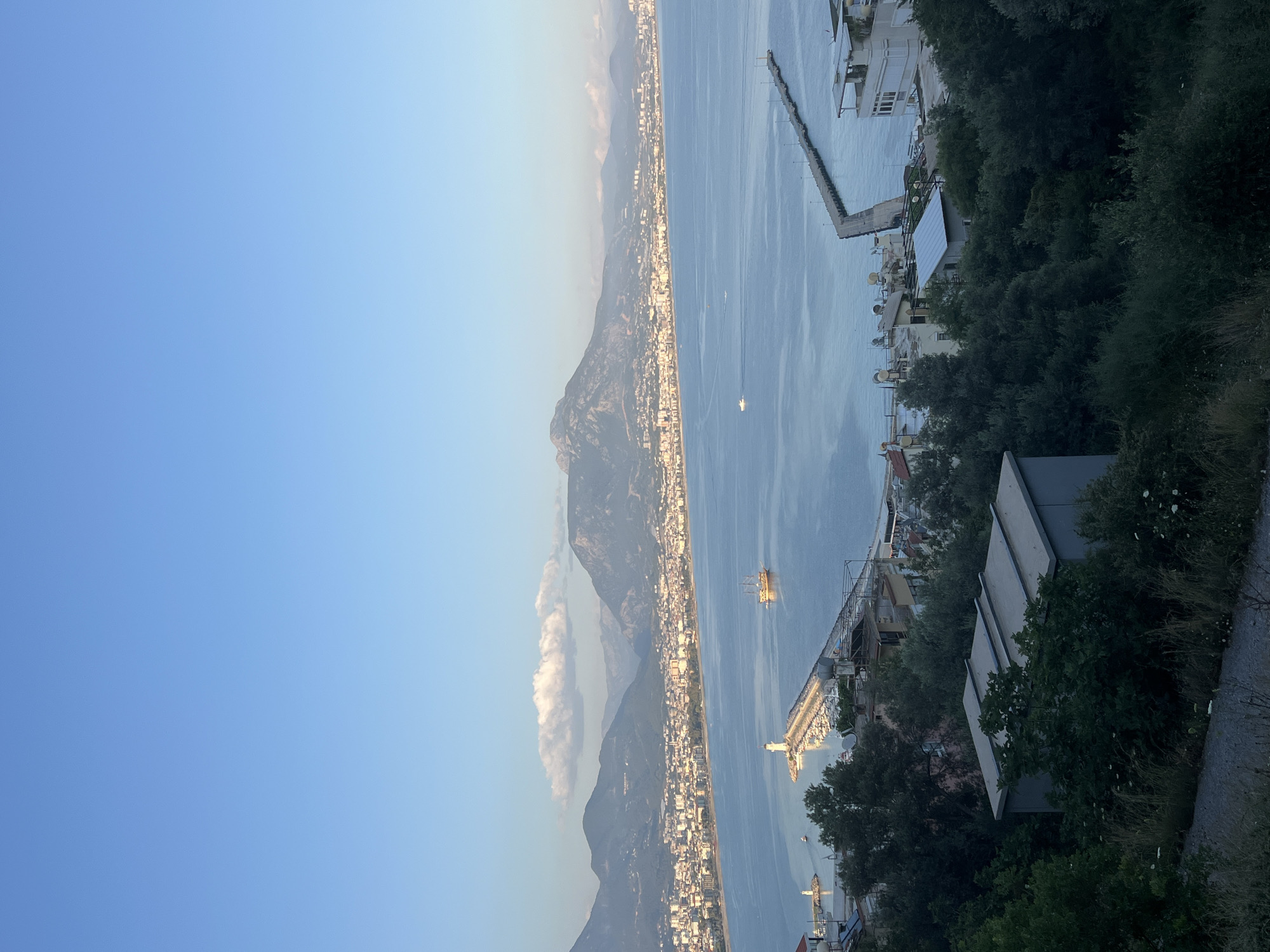 The image captures a high-angle view of Alanya, Turkey. The Mediterranean Sea stretches into the distance, dotted with a few boats. A long pier extends into the sea, flanked by buildings along the coastline. To the left, mountains rise behind the city, partly obscured by haze. The sky is a clear blue. In the foreground, vegetation and rooftops are visible, suggesting the vantage point is from an elevated position within a residential area.