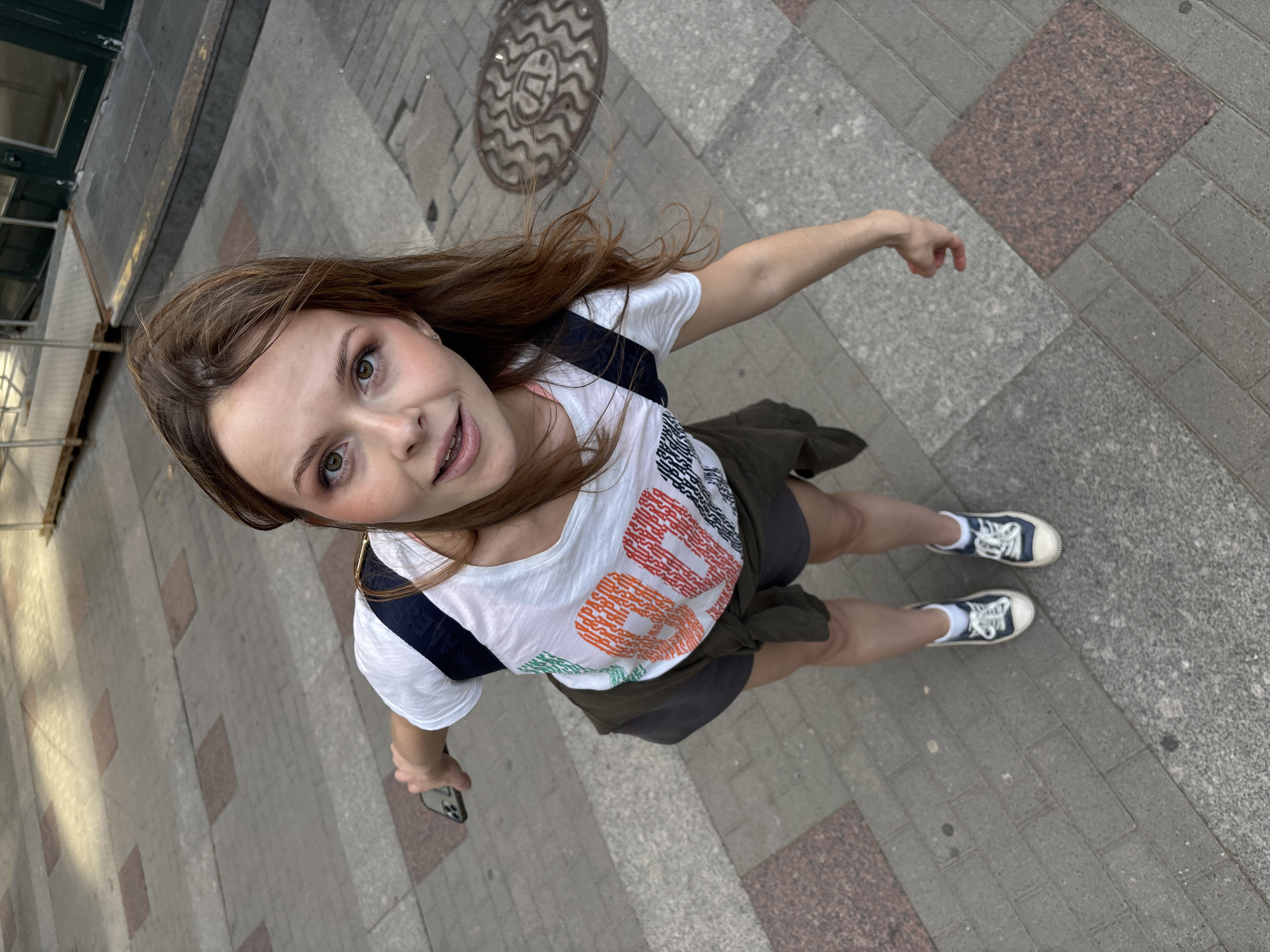 A young adult female is centered in the frame, looking directly upwards at the camera from a low angle. She has brown, shoulder-length hair, a fair complexion, and a slight smile revealing braces on her upper teeth. She wears a white short-sleeved t-shirt with the word "PAUSE" printed across the chest in segmented, multi-colored block letters (green, orange, red, black). A dark olive-green jacket is tied around her waist over dark gray shorts. Her footwear consists of white ankle socks and dark blue canvas sneakers with white laces and toe caps. A dark blue backpack strap is visible on her left shoulder. She holds a dark-cased smartphone in her extended left hand.
The woman stands on a pedestrian walkway composed of alternating gray rectangular paving stones and reddish-brown square granite slabs. To her right (camera's left), a circular metal manhole cover is embedded in the pavement, featuring a wavy pattern and a prominent Cyrillic letter "Д" in its center. This specific design, with the Cyrillic "Д", identifies the location as Saint Petersburg, Russia, typically signifying a storm drain ("Дождевая канализация"). In the background, to her left (camera's right), a dark green building facade with large glass door panels is visible, alongside a yellow curb and a temporary metal ramp leading to the building entrance. The scene is illuminated by ambient daylight.