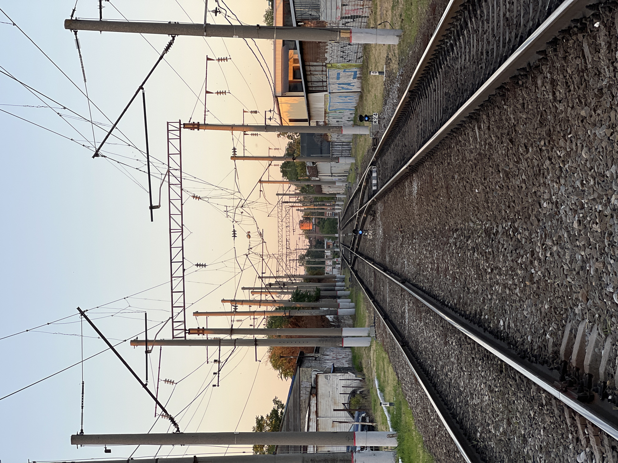 The image depicts a view of a railway track at dusk. The perspective is from the ground, looking along the tracks which diverge into the distance. The sky is a soft gradient of pale blue and peach, indicating sunset. On the left side of the tracks, a series of concrete poles supporting overhead electrical wires for the train system are visible, stretching into the distance. Some of these poles have metal framework attached, and insulators are clearly visible. There are also buildings and structures along the side of the railway, with some showing signs of graffiti. On the right side of the image, the railway track is covered in ballast, with the metal rails gleaming. The scene is likely in a semi-urban or industrial area, given the presence of buildings and industrial infrastructure. The overall mood is calm and serene, characteristic of the end of the day. There is no visible activity or people in the frame. The location is identified as Krasnodar, Russia.