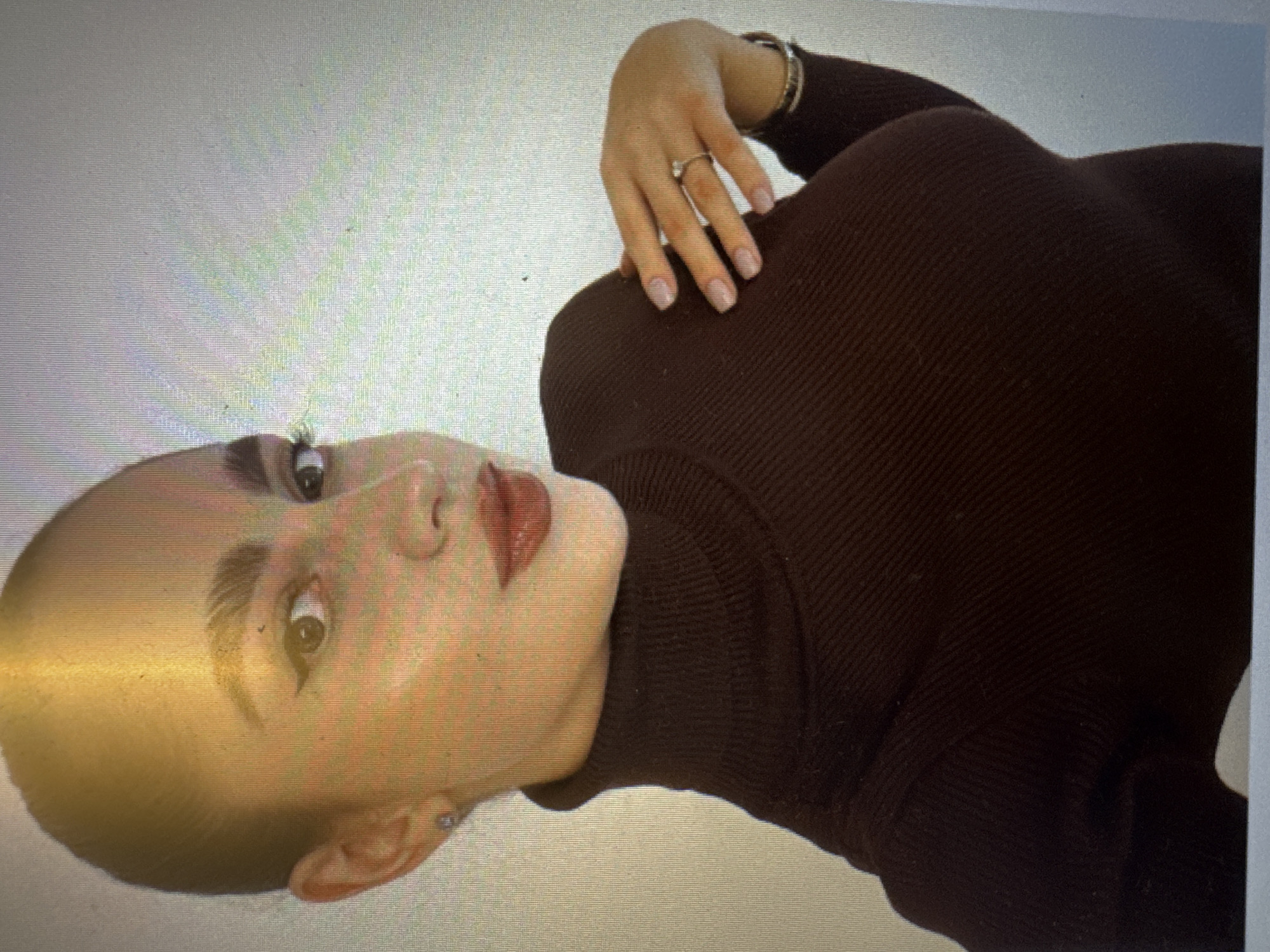 A woman with dark hair pulled back tightly on her head and prominent eyebrows, wearing a dark brown, ribbed, high-necked sweater, poses for a photo. Her left hand is placed on her chest, adorned with a ring on her right ring finger and a bracelet on her right wrist. She wears subtle makeup, including eyeliner and lipstick. The background is a plain, off-white wall. There is no indication of the city of Murcia, Spain, in the image.