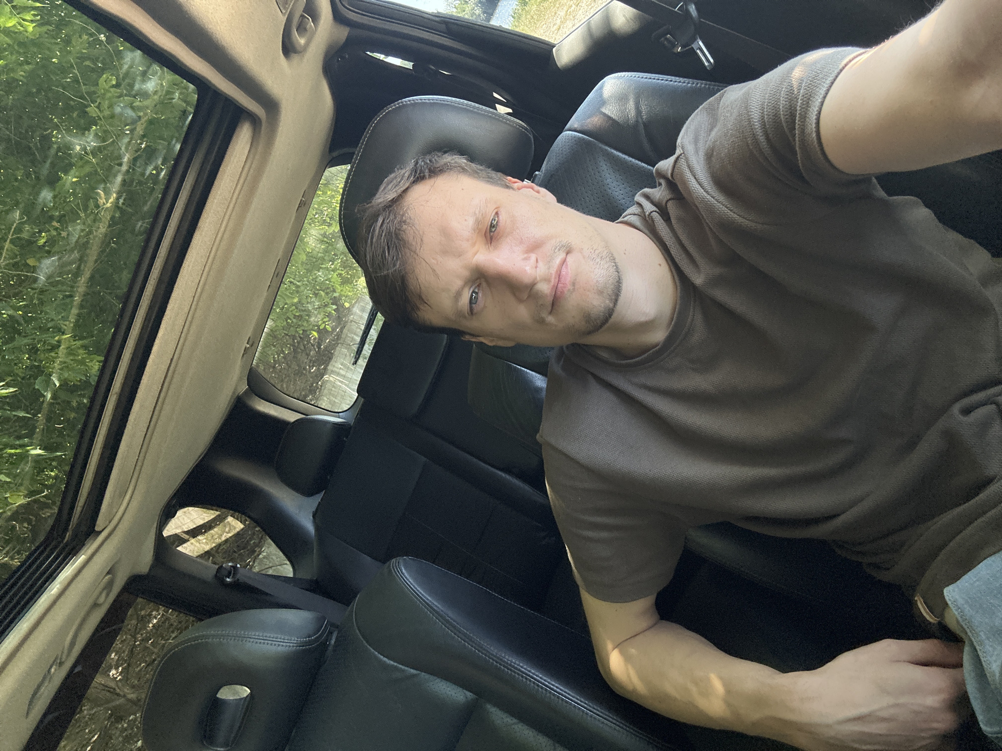 A man is sitting in the driver's seat of a car, looking into the camera. He has short, brown hair, blue eyes, and a light beard. He is wearing a brown t-shirt. The car has a sunroof, and the interior appears to be black leather. Outside the car, there are trees and sunlight. The scene is consistent with being in a car on a sunny day, possibly in a rural or suburban area. The location in Makiv, Ukraine is not discernible from the image itself.