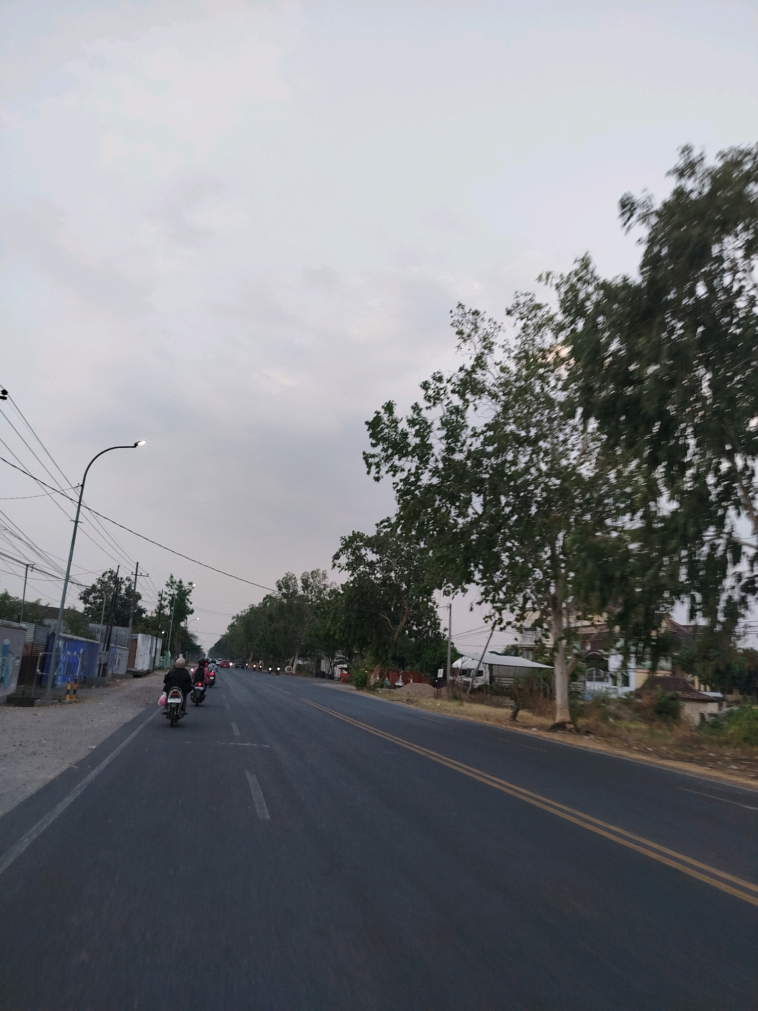 The image captures a street scene in Babadan, Indonesia, during what appears to be dusk or overcast conditions. The asphalt road, marked with double yellow lines, stretches into the distance. Several motorcycles are traveling in the same direction, with riders visible. On the left side of the road, there is a gray sidewalk, a metal fence, and a blue-painted wall with some graffiti. A lamppost with a single light stands tall on the left. Trees line the right side of the road, with one particularly large tree in the foreground obscuring part of the view. Beyond the trees, some buildings are partially visible. The sky is a muted gray, indicating diffused light.
