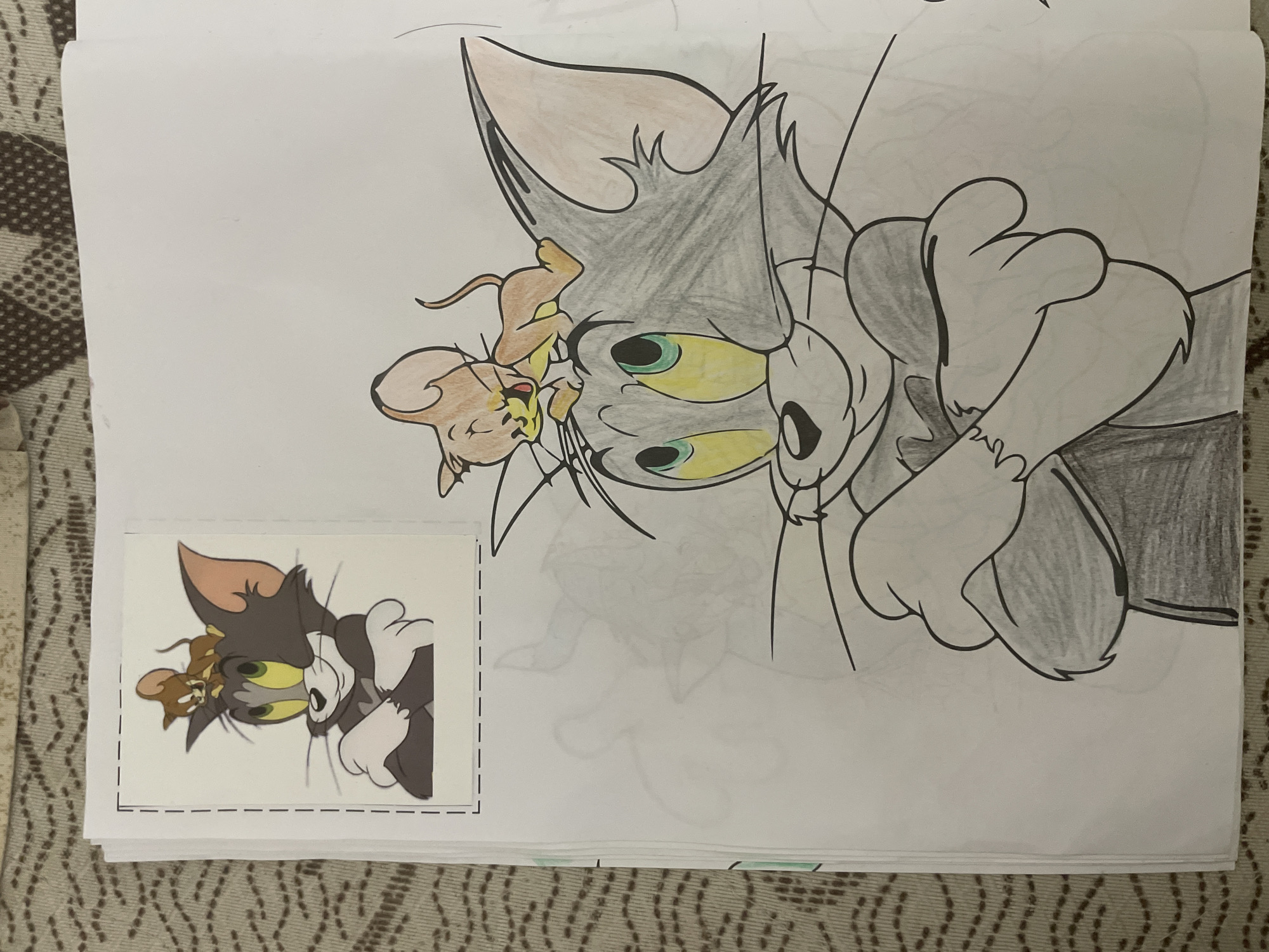 A horizontally oriented drawing of the cartoon characters Tom and Jerry is depicted on a white sheet of paper, partially colored. Tom, the grey cat, is shown with wide, surprised yellow eyes and green irises, with his head and parts of his body colored in shades of grey. His inner ears are a light pinkish-brown. Jerry, the brown mouse, is perched playfully on Tom's head/face, looking towards Tom's nose. Jerry is colored in shades of brown and orange, with pink ears and a reddish mouth/nose area.
In the bottom-left corner of the page, a smaller, fully colored sticker featuring the same image of Tom and Jerry is attached. This sticker is outlined with a dashed line, suggesting it is a cut-out or a guide. The paper with the drawing rests on a patterned surface, which appears to be a light brown or beige rug or blanket with a repeating dark brown geometric design. The drawing seems to be a coloring page that has been partially completed. The scene captures a typical mischievous or playful interaction between the cat and mouse. The image was captured in Ihtiman, Bulgaria.