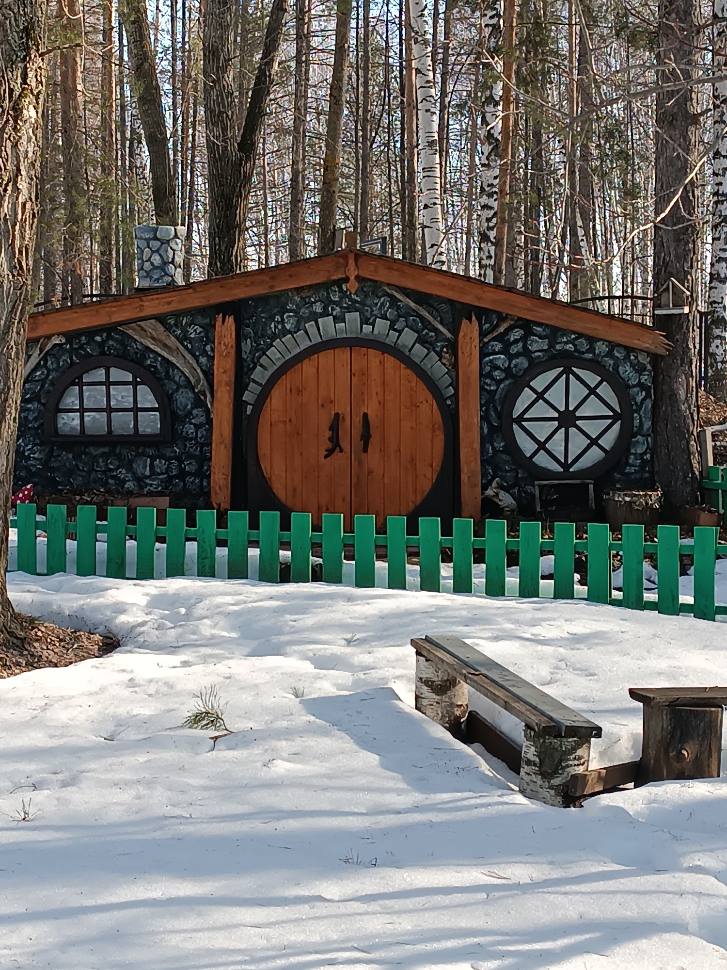 The image shows a whimsical, fantasy-style hobbit-like house situated in a wooded area, possibly a park or recreation area. The house has a round wooden door and two round windows, all set into a stone-like facade. The roof is made of dark wood. A small stone chimney is visible on the roof. A green wooden fence surrounds the front of the structure. In the foreground, there's a wooden bench partially covered in snow, and snow covers the ground. Tall trees, mostly bare of leaves, surround the house. This location is within the city of Chaikovsky, Russia.