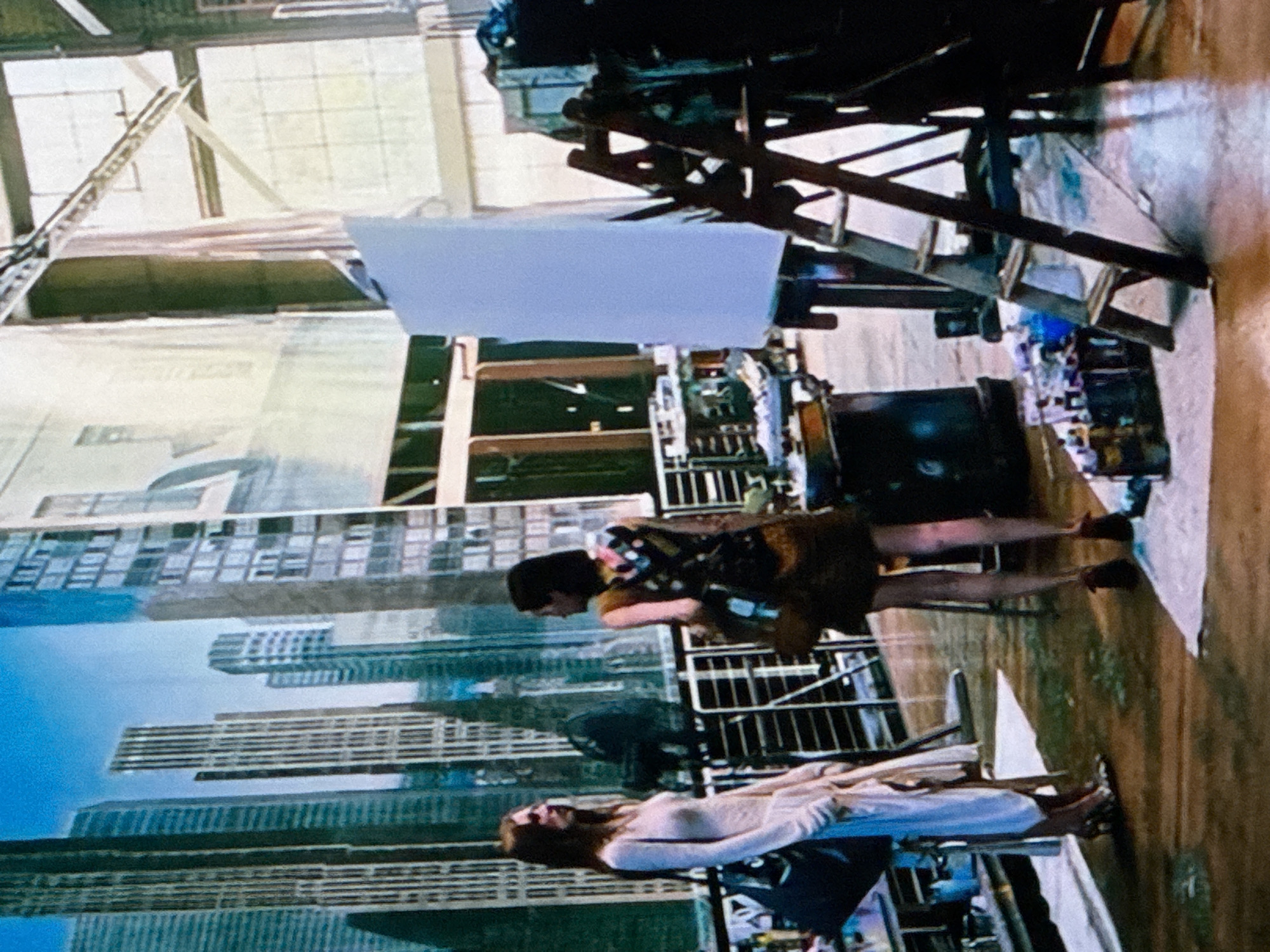 The image shows an indoor scene with two people, one lying on a bed-like structure and the other bending over. In the background, there's a large backdrop featuring a city skyline, possibly of an unknown city due to the "Unknown, Unknown" specification. An easel stands with a white canvas. The environment appears to be a studio or warehouse-like space, as indicated by the exposed metal structure and the wooden floor. Various objects are scattered around the floor.