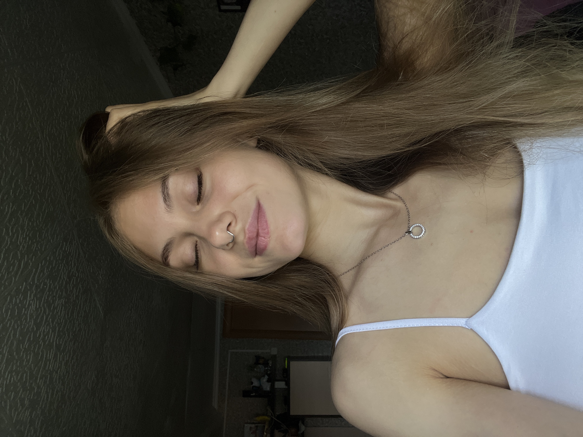 A portrait-style image, rotated 90 degrees clockwise, depicts a young woman with long, light brown hair in an indoor setting in Фоки, Russia. She is shown from the chest up, with her head positioned towards the left of the frame in its current orientation. Her eyes are gently closed, and a subtle, serene smile plays on her lips, conveying a sense of contentment or peace. She wears a silver septum piercing and a delicate silver necklace with a circular pendant. Her attire is a simple white spaghetti strap top. One of her hands is visible near the top of her head, gently touching her hair. The background on the left features a dark, textured wall, while the right side and bottom reveal hints of a room with indistinct objects and reflections, indicating an interior environment. The lighting is soft and diffused. No text is visible in the image.