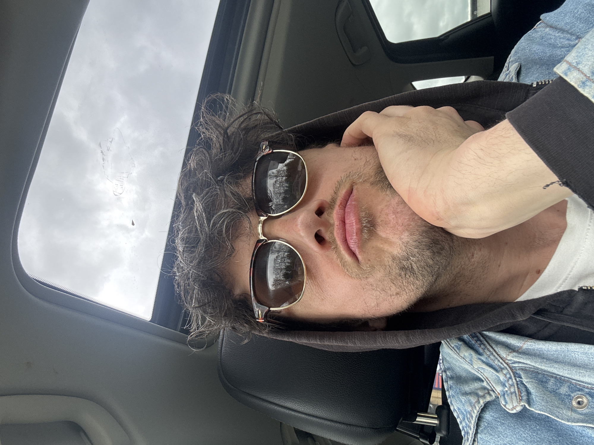 The image depicts a man inside a car, looking upwards towards the open sunroof. He is wearing sunglasses, a hoodie, and a denim jacket. His hand is near his face. The sky is visible through the sunroof. The location, South Kesteven, United Kingdom, cannot be determined from the visual information provided in the image.