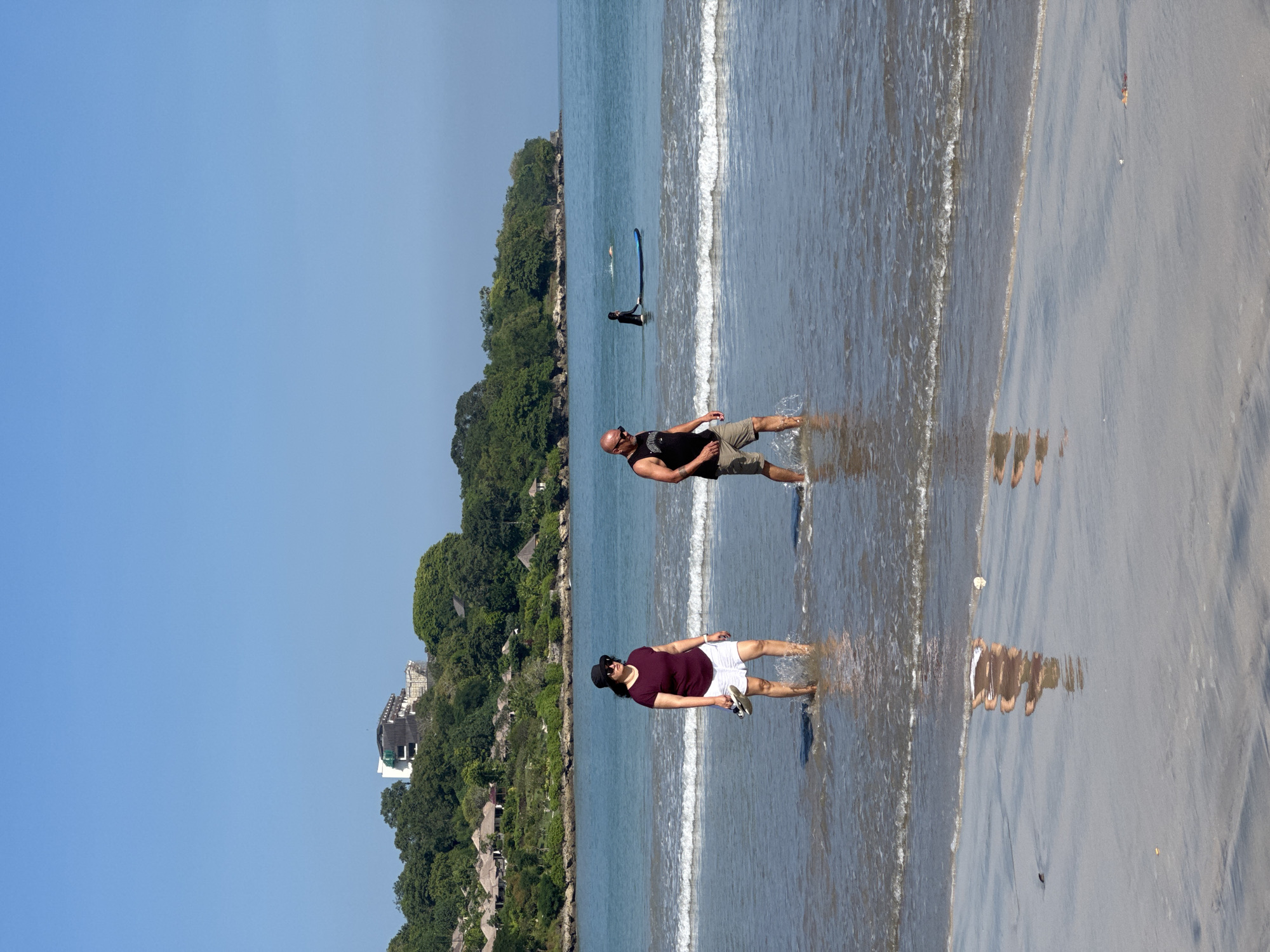 Two people wade in the ocean water at Kuta Selatan, Indonesia, on a sunny day. A man with a shaved head and sunglasses, wearing a black tank top and khaki shorts, is waist-deep in the water. A woman in a black hat, sunglasses, a dark maroon shirt, and white shorts stands next to him, also in the water. In the background, lush green trees line the shore, with glimpses of buildings nestled among the foliage. A surfer stands in the distance. The sand reflects the figures.