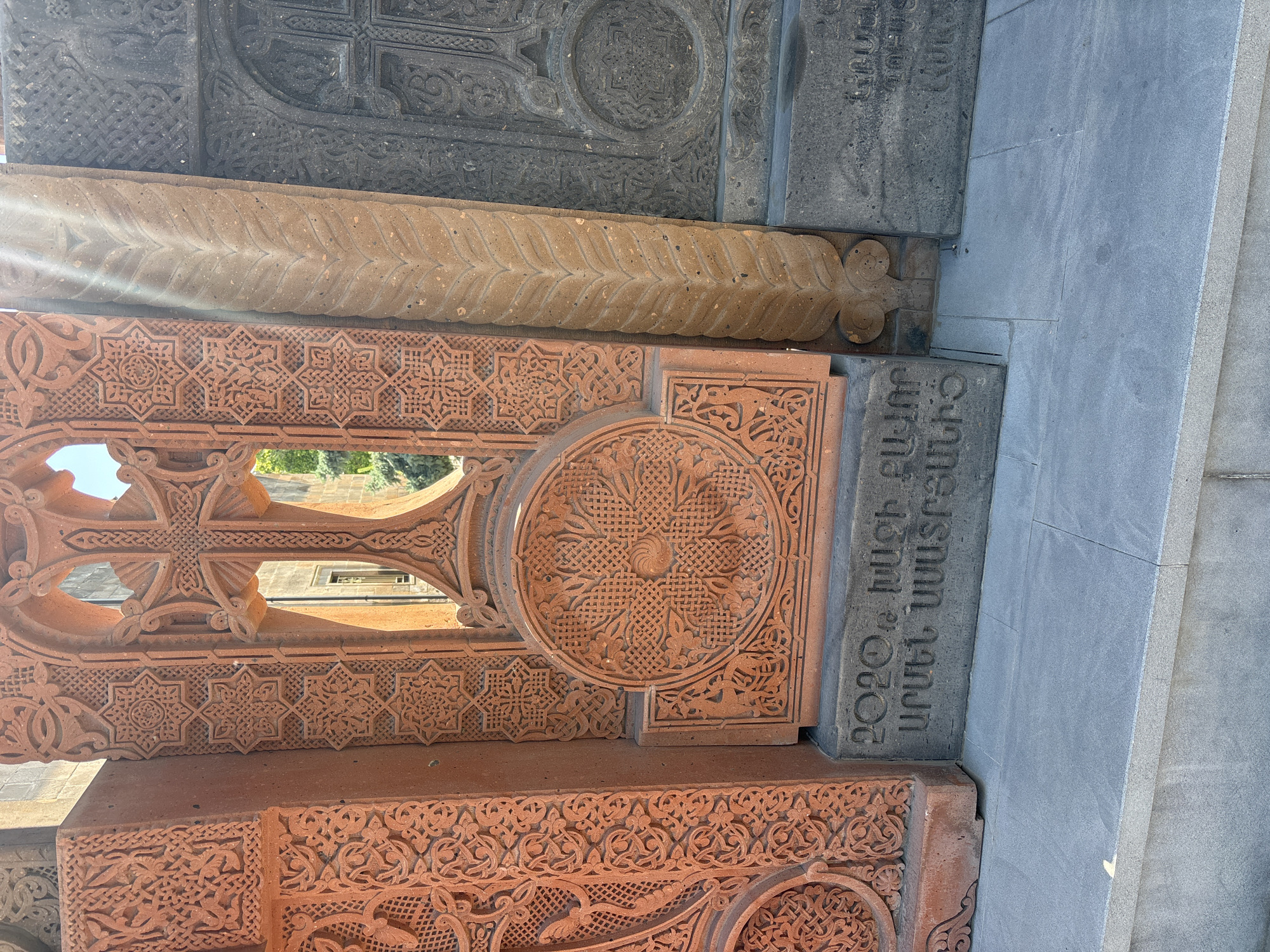 The media features a close-up, angled view of several intricately carved stone slabs, indicative of Armenian Khachkars (cross-stones) or architectural elements. The stones vary in color, including dark grey, light beige, and reddish-brown, and are covered in dense, detailed carvings.
The most prominent feature is a large reddish-brown stone displaying a deeply carved cross adorned with knotwork patterns. Surrounding the cross are geometric star-like designs and continuous floral motifs. A circular rosette, also intricately carved, is visible to the right of the cross. Below this, further elaborate patterns with stylized animal or plant forms are carved into the stone. Above the reddish-brown stone, a light beige slab features a prominent braided or leaf-like pattern. Further above, a dark grey stone is visible with more complex, intertwining geometric and knotwork designs.
The scene is illuminated by bright daylight, with strong sunlight casting shadows that accentuate the three-dimensional depth of the carvings. Through the openings in the carved cross, green foliage and parts of a building structure can be seen in the background, out of focus, suggesting an outdoor setting like a monastery or churchyard.
On a dark grey stone slab towards the lower right of the primary image, Armenian text is clearly visible. The inscription reads: "2020թ. ՄԱՐՏԻ 15ԻՆ ԱՐԱՐԱՏԻ ՄԱՐԶԻՑ". This translates to "2020, March 15th, from Ararat Province," likely indicating the date of creation or dedication of the stone and its place of origin. The image captures the rich artistry and historical detail of Armenian stone carving.