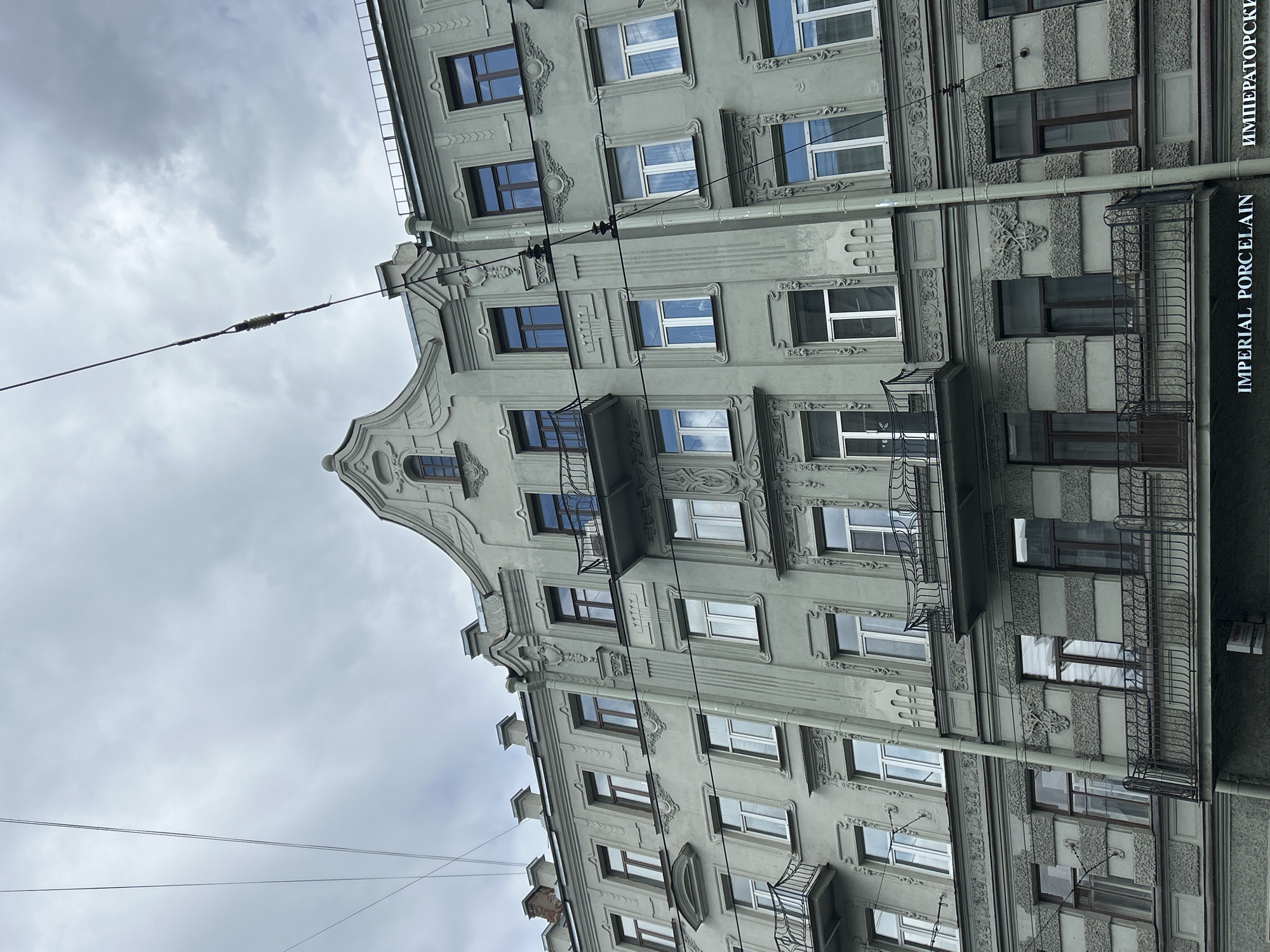 The image shows a close-up, low-angle view of a grey, multi-story apartment building with ornate architectural details. The building features multiple rows of windows, some with dark frames and others with white frames, and several balconies with intricate wrought-iron railings. Decorative elements such as relief carvings and molding are visible on the facade. Overhead, electrical wires crisscross the sky, which is overcast with grey clouds. In the bottom right corner of the image, text is visible, including "IMPERIAL PORCELAIN" and its Russian equivalent, "ИМПЕРАТОРСКИЙ".