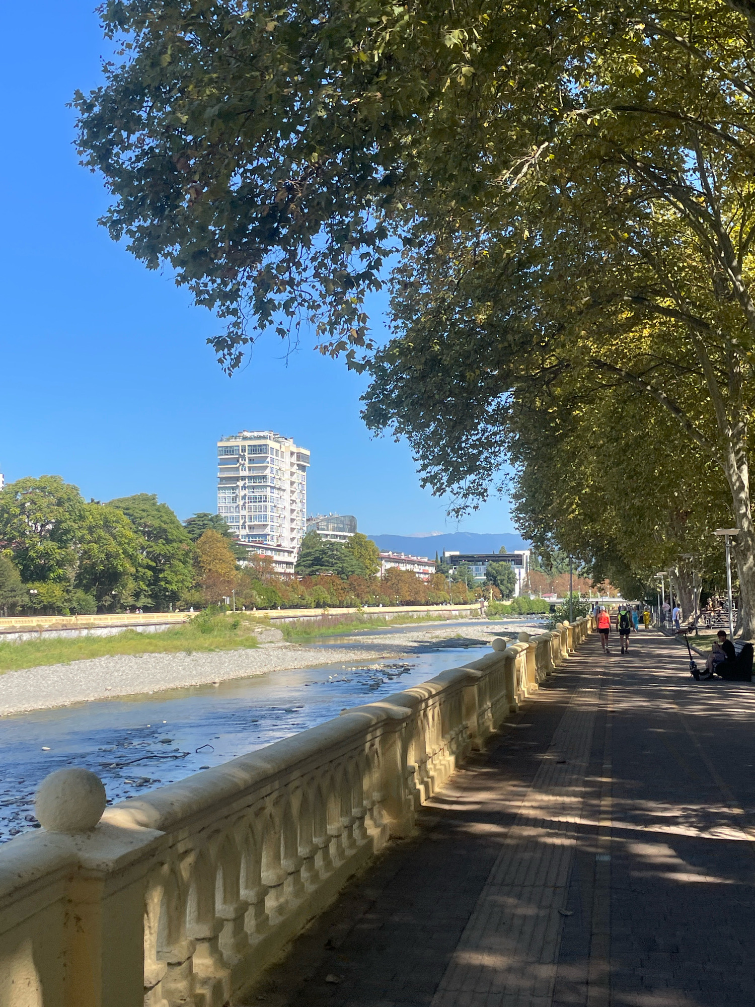 This image depicts a sunny day along a paved walkway adjacent to a river in Sochi, Russia. The walkway is lined with a decorative stone railing and bordered by a dense canopy of mature trees with green leaves, casting dappled shadows on the path. Several people are visible on the walkway; two individuals are running, others are walking, and one person is seated on a bench with an electric scooter nearby. Across the river, the opposite bank features a rocky, shallow riverbed, with lush green trees and a prominent multi-story white building in the background. In the distance, mountains are visible under a clear blue sky. This scene is likely the Reka Sochi embankment in the city of Sochi.