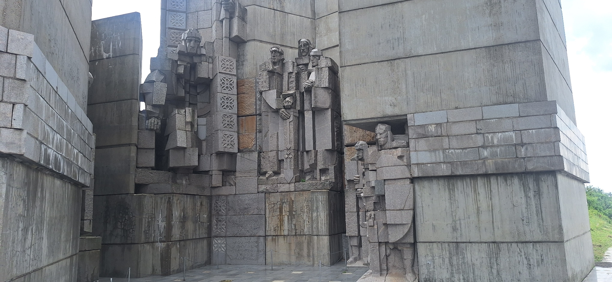 The image shows the Monument to the Founders of the Bulgarian State in Shumen, Bulgaria. The monument is constructed from concrete and stone, with large geometric shapes and carved figures. In the foreground, on the right side of the image, there are two large stone figures standing in an alcove. The figure on the right is larger and wears a shield. Both figures are stylized and appear to be warriors or historical figures. To the left, the monument features several more carved figures that are partially obscured by the concrete structure. These figures are also stylized, with geometric patterns incorporated into their design. The monument is surrounded by a paved area and a grassy slope is visible in the background on the far right. The sky is overcast.