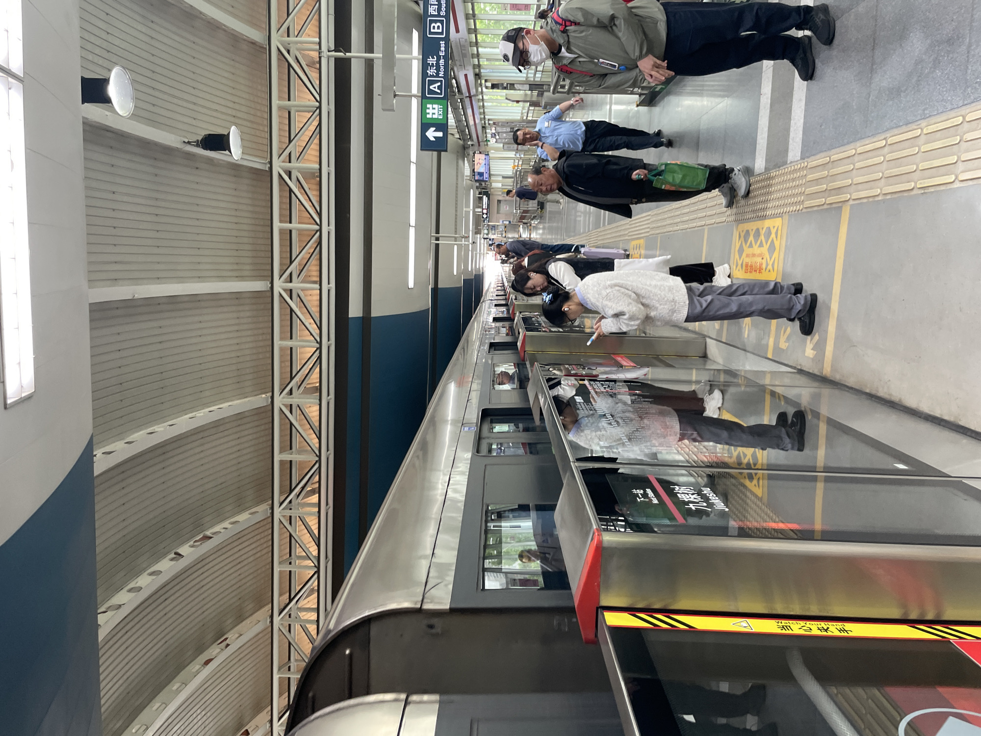 The image depicts a subway station platform in the Tongzhou District of China. Passengers are lined up behind a safety line of tactile paving and yellow warning markers, awaiting a train. Some individuals are looking at their phones. Two uniformed personnel are seen at the far end of the platform, one wearing a mask. Overhead signs indicate exits labeled "A" (North-East) and "B" (South-West). The station has a high, arched ceiling with visible support beams and lighting fixtures. The train doors are visible, reflecting the platform and passengers. A warning sign in Chinese and English "Watch Your Hand" is visible on the train doors.