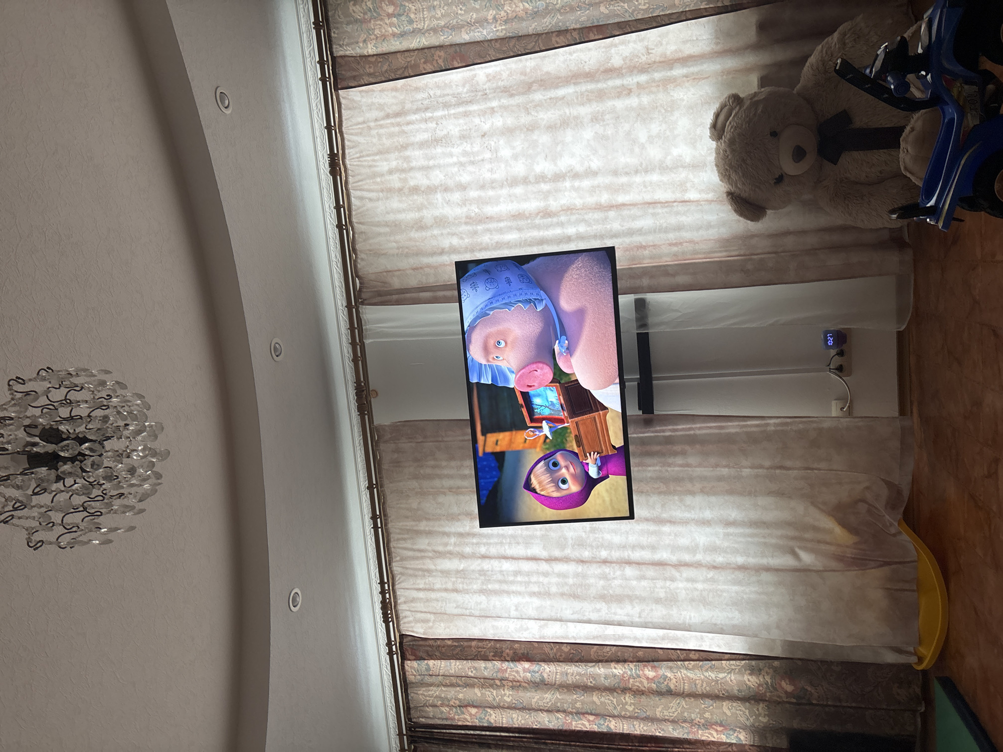 A vertically oriented indoor shot shows a room in Ferma, Russia, with a white arched ceiling on the left featuring a chandelier and recessed lights. A television is mounted on the wall, displaying an animated image of a pig and a young girl (Masha and the Bear). To the right of the TV, a beige teddy bear sits next to a blue child's four-wheeler toy. The wall is covered with curtains that have a beige patterned top and a semi-transparent white middle section. On the floor to the right is a yellow plastic toy.