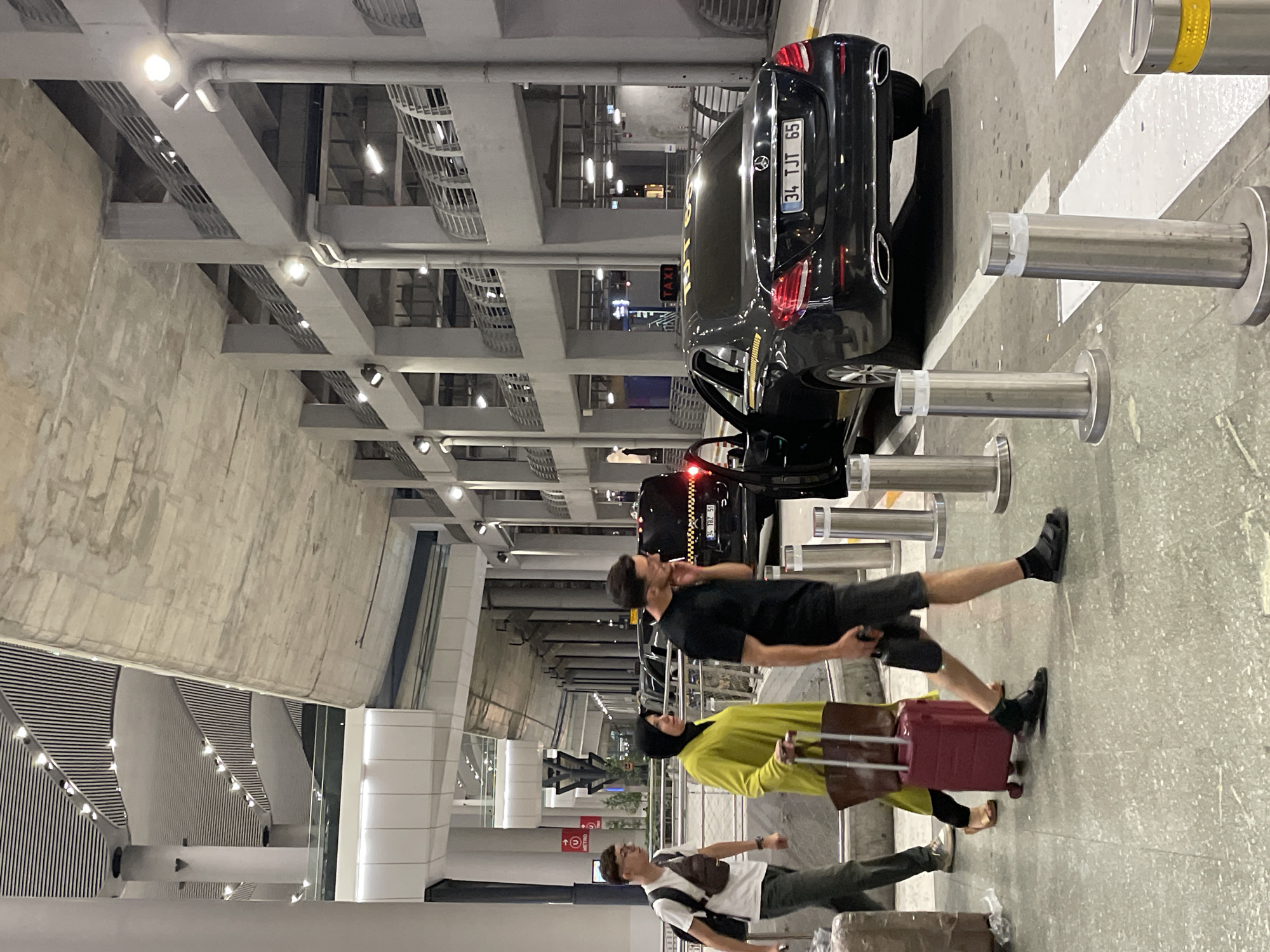 This is a full-frame, eye-level, medium shot of a scene outside an airport terminal in Arnavutköy, Turkey. A group of three people are walking on a pedestrian pathway next to a black Mercedes-Benz sedan with a taxi sign on top. The man on the left wears a white t-shirt, dark pants, and a crossbody bag. The woman in the middle wears a bright yellow headscarf and abaya, and is pulling a red suitcase with a brown tote bag. The man on the right wears a black t-shirt, dark shorts, and sandals. He is holding a black smartphone to his ear. The taxi has the license plate "34 TJT 65". The airport terminal features a modern architectural design with exposed concrete structures and a layered ceiling with integrated lighting. Several metal bollards are positioned along the pedestrian pathway. The lighting suggests it is evening or night.