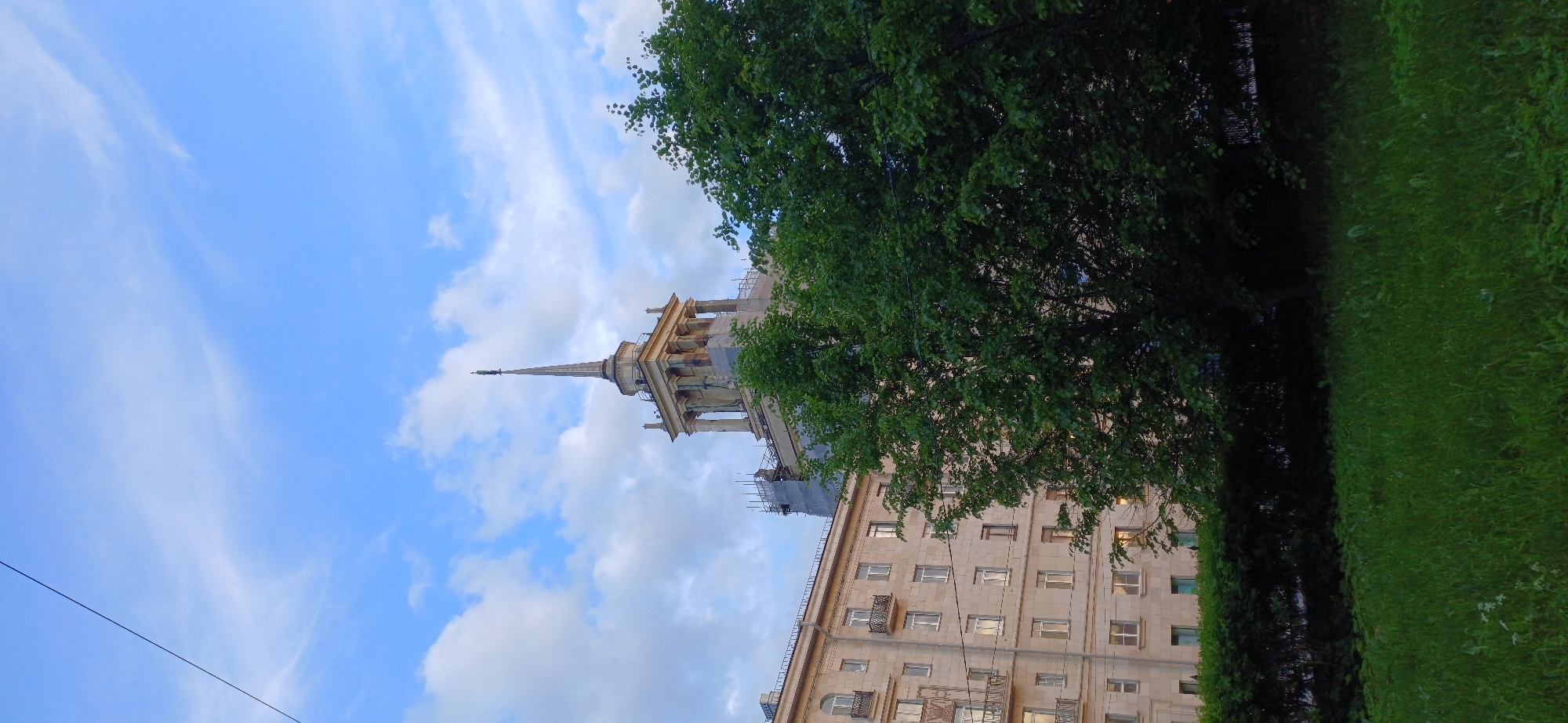 This vertically oriented image captures a bright daytime scene in Saint Petersburg, Russia. The upper and left portions are dominated by a clear blue sky with scattered white clouds, some wispy, others puffier. A prominent architectural landmark, a tall building topped with a slender, ornate spire, rises towards the sky. This building features classical elements, including columns and pediments, and appears to be undergoing renovation, indicated by visible scaffolding on its upper sections.
To the right and below the spired building, a multi-story, light-colored building with numerous rectangular windows and some small balconies is visible. Its facade suggests a mid-20th-century architectural style, possibly Stalinist or neoclassical. A large, leafy green tree partially obscures the view of the buildings, casting some shade and adding natural color to the scene. Below the tree, a strip of vibrant green grass extends across the foreground. A thin, dark line, possibly a power cable, stretches across the lower left of the sky. The overall impression is one of a typical urban landscape, serene under a pleasant, partly cloudy sky, with no specific activities or people visible. There is no visible text.