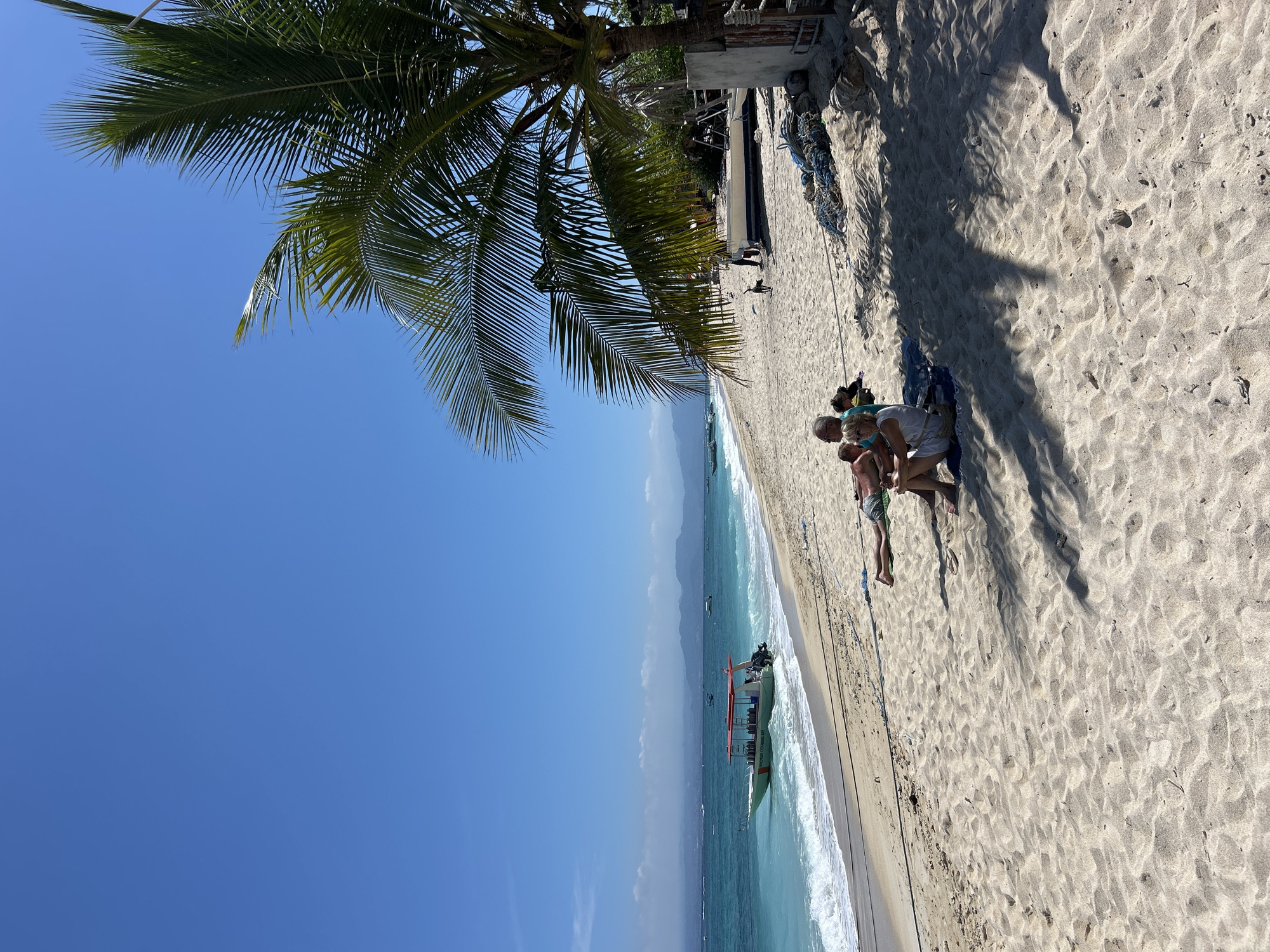 A couple relaxes on a sandy beach in Nusa Penida, Indonesia, under the shade of a palm tree. The man, wearing green shorts, lies on a towel, while the woman, in a white dress, sits beside him, leaning her head on his shoulder. In the background, turquoise water laps against the shore, and a dive boat is anchored near the beach. The sky is a clear blue, with distant mountains visible on the horizon. Several other people are scattered on the beach, and a dog is seen walking in the distance. Ropes and fishing nets are piled near a small structure on the right side of the image.