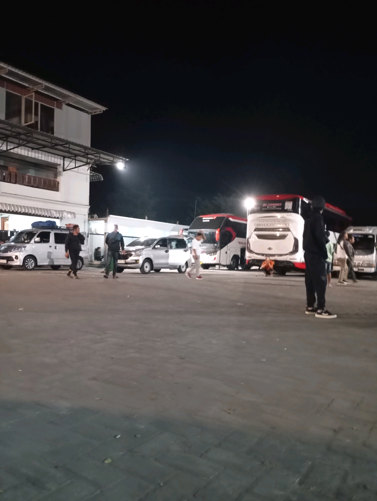 In the city of Purworejo, Indonesia, an outdoor scene unfolds at night, illuminated by artificial lights. The foreground is a paved area, transitioning from a lighter gray to a darker gray with a brick pattern. In the middle ground, several vehicles are parked: a white van on the left, a silver car and another van to its right, and two large buses further back. The buses are white with red accents and appear to be intercity coaches. A building with a metal awning and windows is visible on the far left.
Several individuals are present in the scene. Two men in dark clothing stand near the white van on the left, one walking away from the viewer and the other facing towards the viewer. Another man in dark clothing stands on the right side of the frame, facing away from the viewer and towards the buses. A child in a white shirt and light-colored pants is visible near the silver car, walking towards the right. Another person in a white shirt and dark pants is walking away from the viewer on the far left, near the white van. Additionally, a few individuals are standing near the buses, some appear to be in light-colored clothing. The overall atmosphere suggests a transportation hub or rest stop at night.