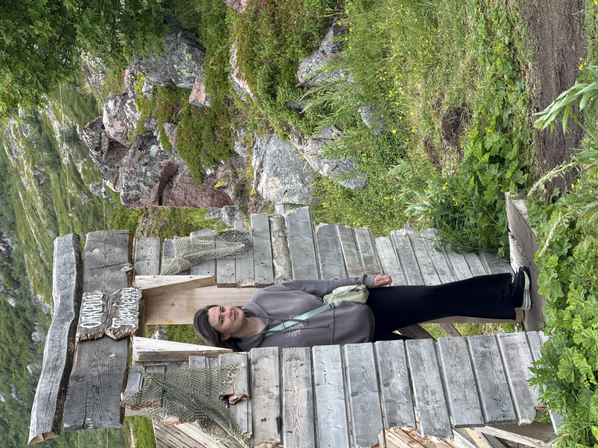 A woman stands in the doorway of a rustic wooden structure in Teriberka, Russia. The structure appears to be made of weathered, uneven planks, with a sign above the entrance that reads "Откроем Теребирь" (We will open Terebir). Fishing nets are draped over parts of the wooden frame. The woman is wearing a grey hooded sweatshirt, black pants, and sneakers. She has a small, light-colored crossbody bag. The background shows a rocky, green, and hilly landscape, typical of the Arctic coastal environment of Teriberka. There are scattered rocks, dense green vegetation, and some small yellow flowers in the grass. The sky is overcast.