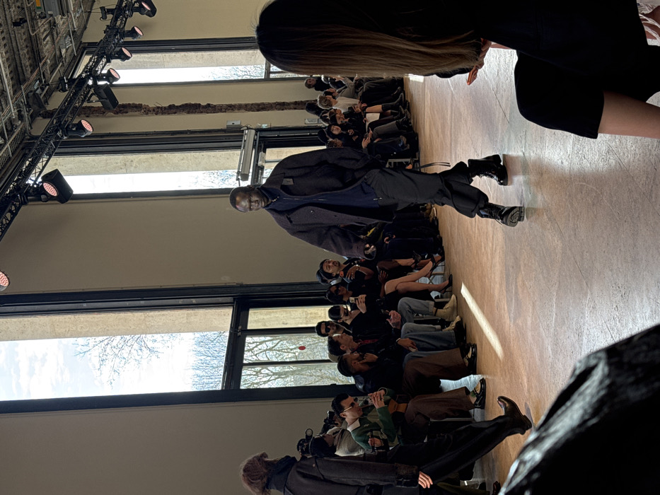 An indoor fashion runway event is depicted within an unidentifiable event hall in Unknown, Unknown. A dark-skinned, bald male model, wearing a dark blue collared zip-up jacket over a dark blue top, dark baggy trousers, and black combat boots, walks from left to right on a light brown floor, maintaining a direct gaze. On the far left, a second model with reddish-brown hair and dark clothing is partially visible, walking away from the viewer. A seated audience occupies rows along the left side of the runway, observing the models; multiple individuals are holding smartphones, suggesting active photo or video capture. The venue features high ceilings with exposed metal trusses supporting theatrical lighting fixtures. Large, multi-pane windows line the rear and left walls, revealing bare trees against a bright, cloudy sky, indicating an outdoor, natural environment. The interior walls are light beige.