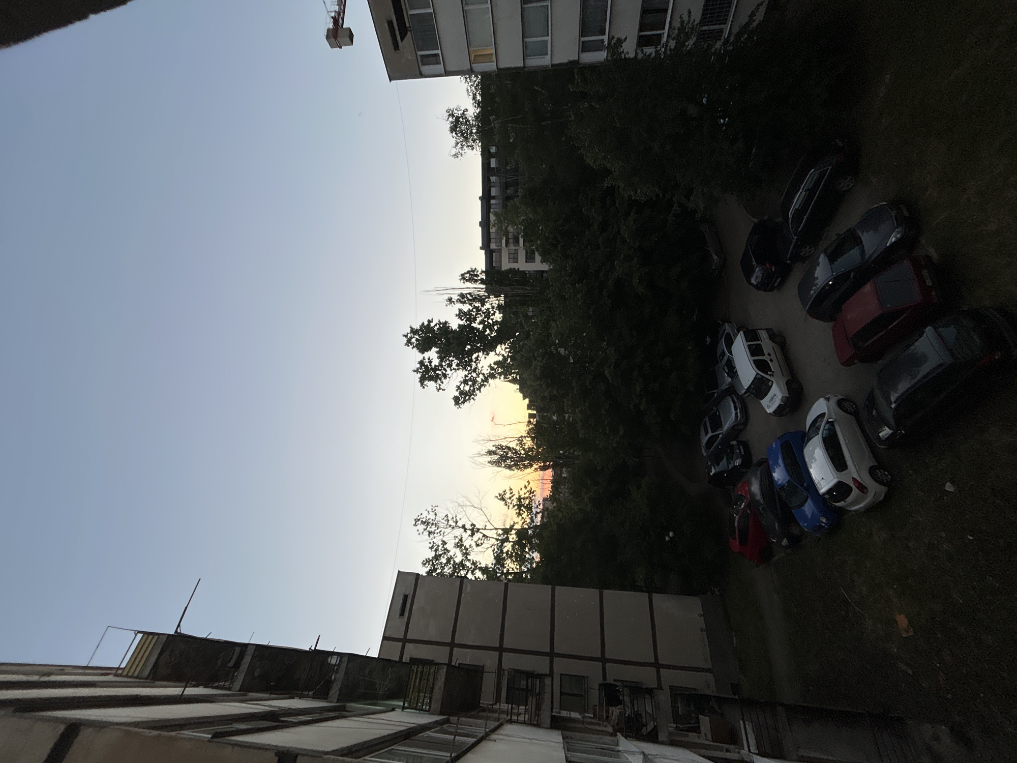 The image depicts a dusk scene in an urban environment, likely Sofia, Bulgaria. A building with a flat roof dominates the foreground. Above and to the left, the sky is a pale blue-grey, indicating twilight. A cluster of trees in the middle ground partially obscures a sunset with shades of orange and red. Several parked cars are visible in the courtyard area, including cars of red, blue and white. A construction crane can be seen in the upper left corner, near a taller building with visible windows. The overall environment suggests a residential area in Sofia, marked by typical urban infrastructure like apartment blocks and parking spaces. There are no people visible in the image.