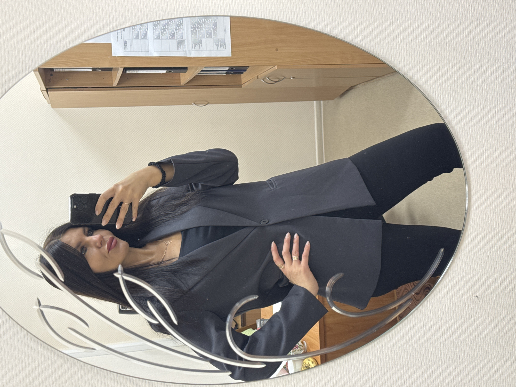 An adult female is depicted taking a mirror selfie, captured from the waist up. The image itself is rotated counter-clockwise. She has long, dark hair and is dressed in a black blazer over a dark top, with dark pants. She wears a black beaded bracelet on her left wrist, multiple rings on her right hand, and a gold necklace featuring a small cross pendant. She holds a black smartphone in her left hand, positioned to capture her reflection. Her expression is a slight pout with parted lips.
The mirror is circular and features decorative, silvery, etched or applied floral/leaf-like patterns on its lower left quadrant. Reflected in the mirror, the background reveals light-colored, textured wallpaper, likely off-white or beige. Above the woman's reflection, a wooden shelf or desk unit is visible. On this unit, several papers or documents with small, illegible text are arranged, along with what appear to be black binders or folders. The setting suggests an indoor room, possibly a personal space, in Chaikovsky, Russia, with lighting indicative of daytime.