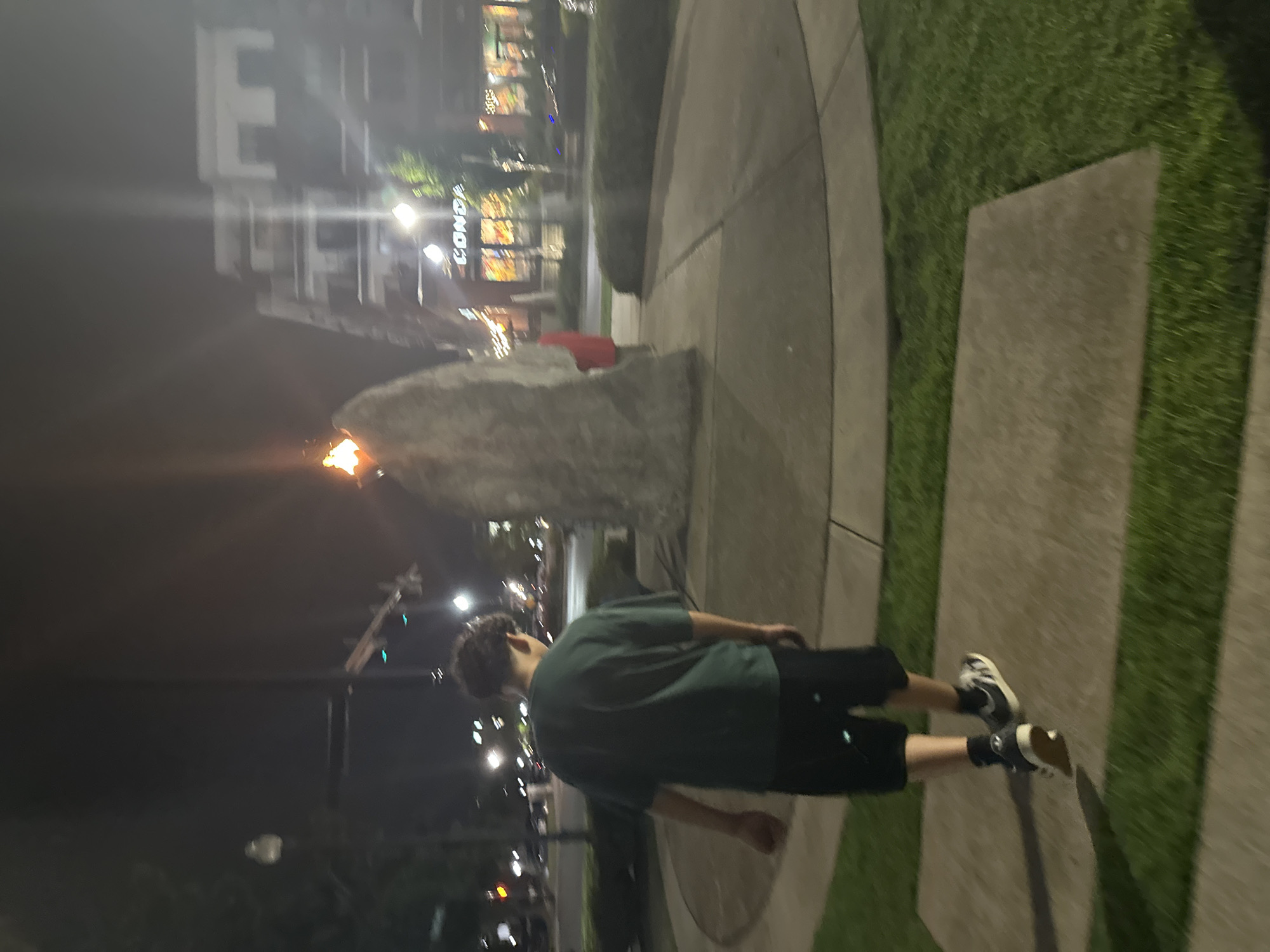 This is a medium shot, taken at night, of a young male walking away from the camera on a paved path. He is wearing a dark green t-shirt, black shorts, dark socks, and white sneakers with black stripes. The man is walking on a concrete sidewalk that is bordered by grass. In the background, there is a large, rough-surfaced stone sculpture with a flame at its peak. Behind the sculpture, there are buildings with illuminated windows, a street with traffic lights, and trees. The overall scene appears to be an outdoor public space, potentially a park or plaza, in Huntsville, Alabama.