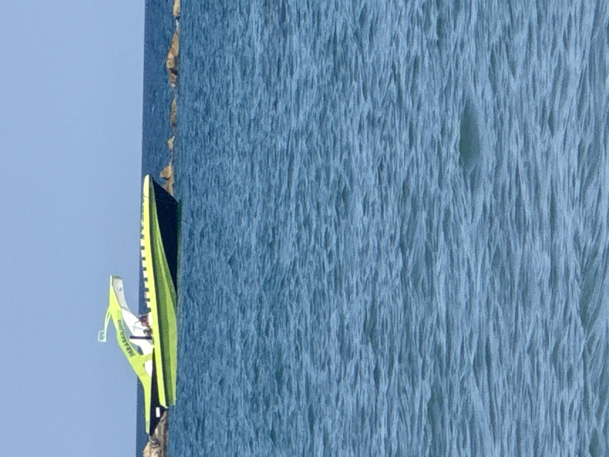 A bright green and black speedboat with a white canopy is visible on the blue sea. The boat is identified by the text "PARASAILING" in white lettering on its side. The horizon separates the sea from a clear blue sky. In the background, a few rocks are breaking the surface of the water. The overall scene suggests a bright, sunny day. The image is located in the Agios Tychona Community, Cyprus.
