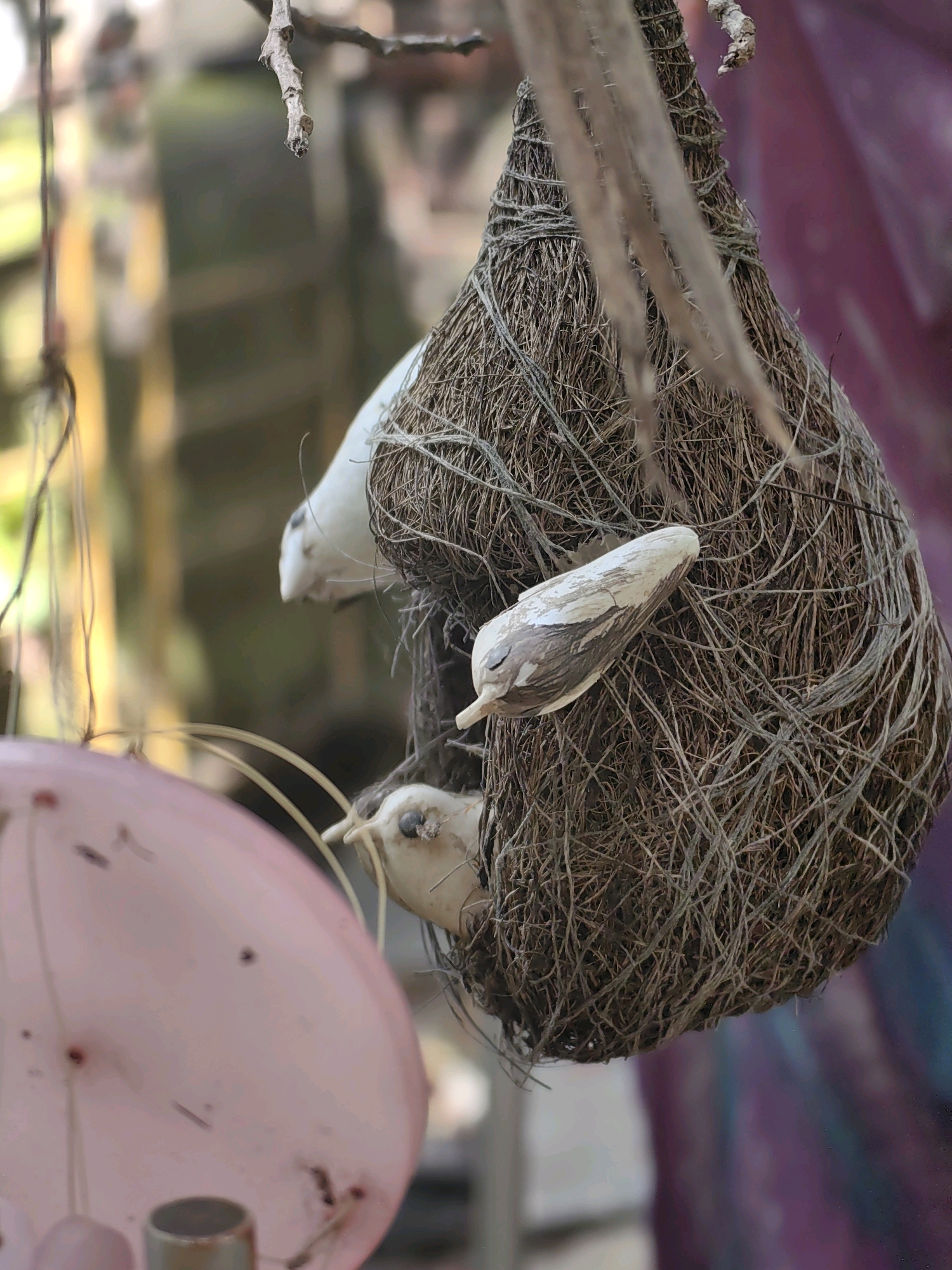A close-up, eye-level shot captures a woven bird's nest suspended by a branch. The nest, primarily constructed of dry grass and thin fibers, has an opening on its underside. Three small, white decorative bird figures are affixed to the exterior of the nest. The foreground features a blurred, pink, semi-circular object, possibly a balloon, with faint dark markings. The background is out of focus, showing muted colors and indistinct shapes that suggest an outdoor environment. The image is taken in Mumbai, India.