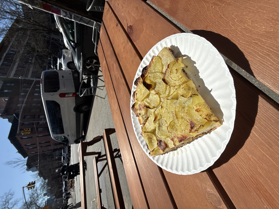 A slice of savory potato tart, topped with thinly sliced baked potatoes, is presented on a white fluted paper plate on a brown wooden slatted outdoor table. The tart exhibits golden-brown edges on some potato slices and a visible pastry base. Strong sunlight casts distinct shadows from the plate onto the table. In the immediate background to the left, an empty, identical wooden picnic table stands on a sidewalk. Beyond the sidewalk, a street scene unfolds in a district of New York City. A white commercial-style van is parked on the street, followed by a dark-colored sedan. A motorcyclist in dark attire and a helmet is visible further down the street on the left. Multi-story brick buildings, characteristic of New York architecture, line the street, featuring fire escapes. Bare trees are present, suggesting a late winter or early spring setting. A traffic light signal is visible at an intersection in the upper left background. The sky is clear and blue.