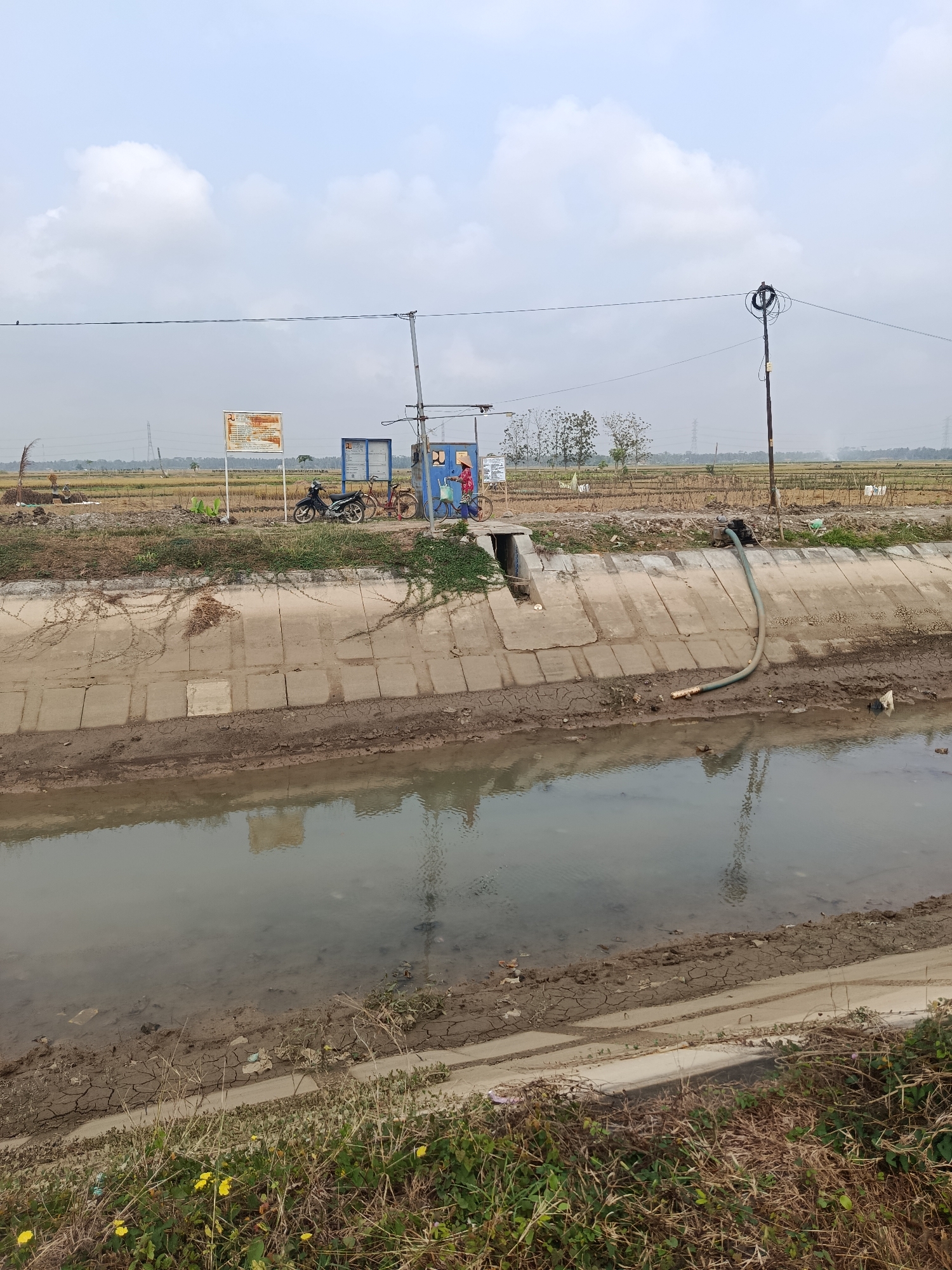 A canal with muddy, cracked earth banks is in the foreground, reflecting a cloudy sky. On the far bank, a concrete structure lines the canal. Above the concrete, dry, brown vegetation grows, with a few scattered yellow flowers in the lower right corner. Further back, a field of harvested crops is visible. Several objects are present: a blue electrical box with a metal pole and wires attached, a motor with a hose leading into the canal, two bicycles, and a motorcycle. Two individuals are near the bicycles, one wearing a red hat and carrying a blue bag, and another wearing a yellow hat. A large sign with text is also visible behind the vehicles. Power lines stretch across the sky. The scene appears to be in a rural or agricultural area, possibly near Bantarsari, Indonesia, given the context.