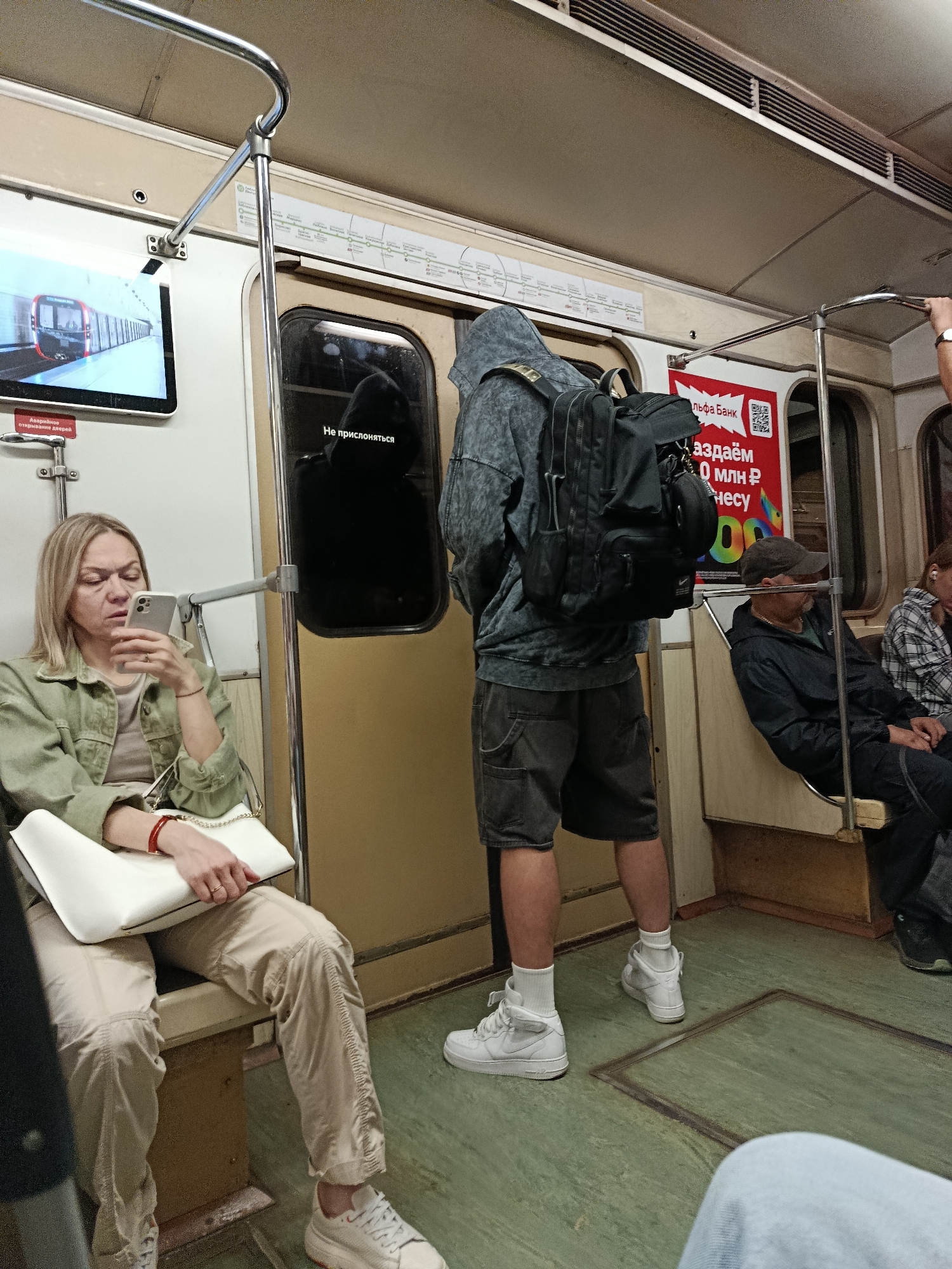 Inside a Moscow Metro car, a woman in a green jacket and beige pants sits on a bench, engrossed in her smartphone. Across the aisle, a person wearing a hooded sweatshirt and shorts, carrying a large backpack, stands near the door. Several other passengers are visible, including a man in a dark jacket and cap to the right, and a person in a plaid shirt further back. Advertisements are displayed on the walls, including a "Qiwi Bank" promotion. The car's interior features metallic poles, handrails, and worn seating. A screen on the left displays a train on a track. The floor is a greenish color with a visible grate.