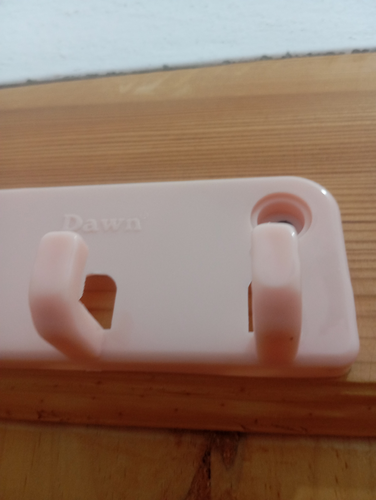 A light pink plastic hook rack with the embossed word "Dawn" is mounted on a wooden surface. The rack has two hooks extending outwards, one on the left and one on the right. The background consists of a white wall above a wooden surface. The image is an indoor, close-up shot of the hook rack.
