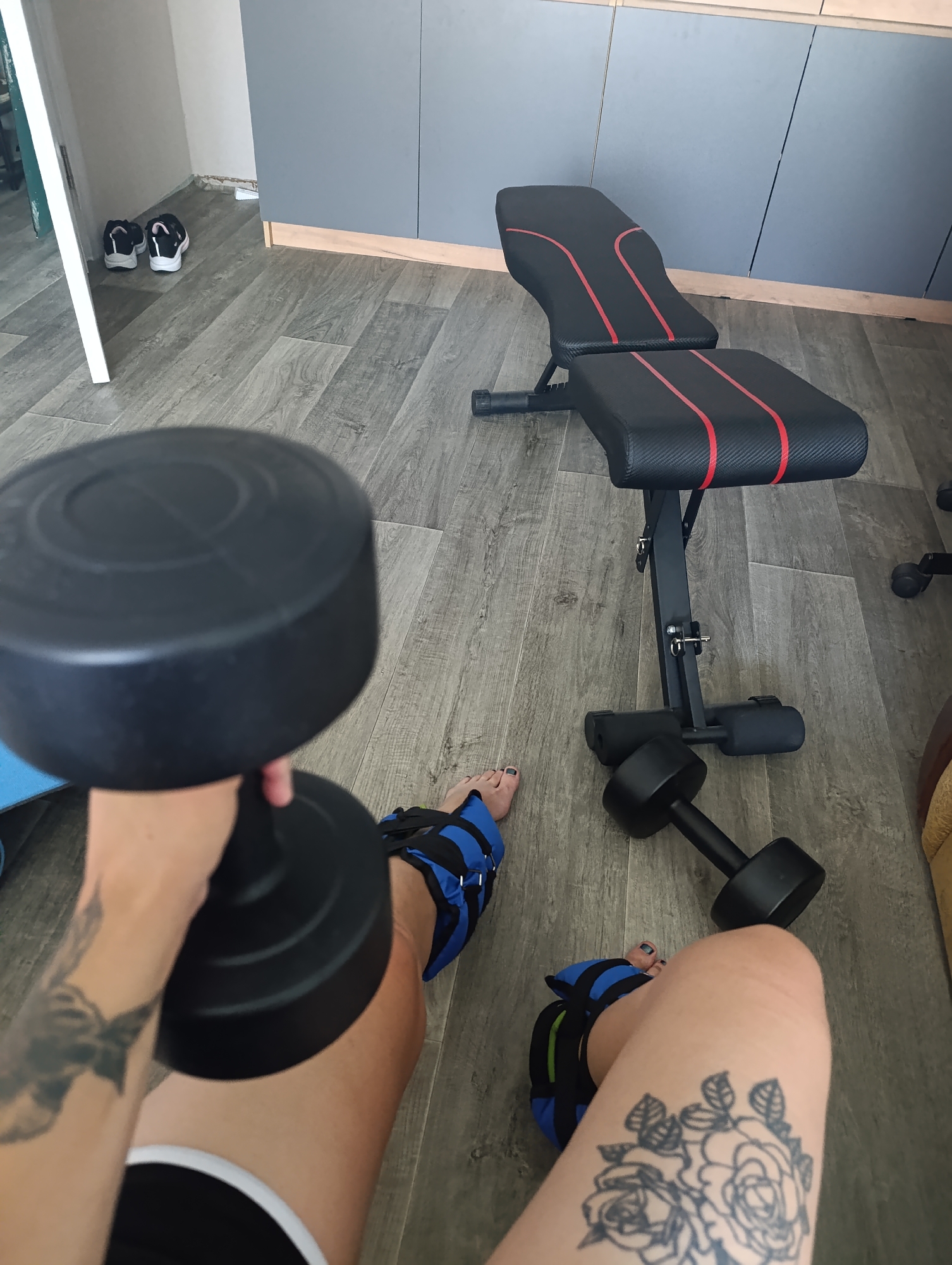 The image shows a home gym setting. A person is sitting on the floor, holding a dumbbell. They are wearing ankle weights and black shorts. Next to them are other dumbbells and a weight bench. In the background, there are two pairs of shoes and a grey cabinet. The floor is made of wooden planks. The scene is indoors, likely in an apartment or house. The context suggests a workout session. The location is specified as Novokuybyshevsk, Russia.