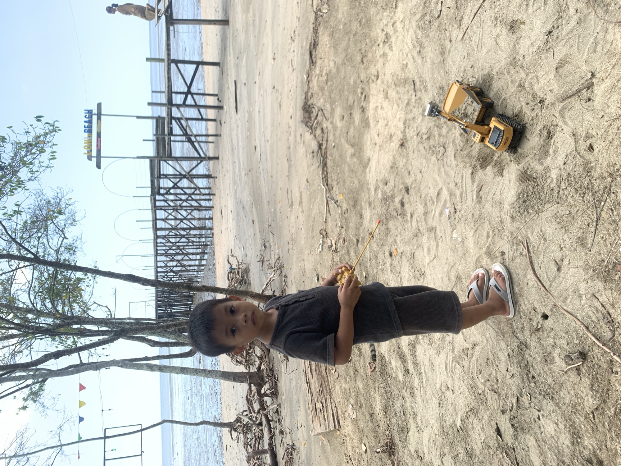 A young boy stands on the sandy beach in Tanjung Limau, Indonesia, wearing a dark-colored outfit and white sandals. He holds a yellow object in his hands. A yellow toy excavator sits on the sand to his right. Behind him, trees line the beach, and a pier structure with a sign reading "ALOW BEACH" is visible. The ocean is in the background under a bright sky.