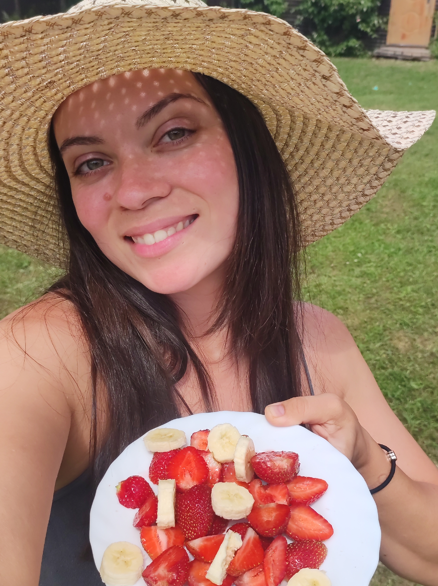A woman wearing a straw hat is holding a white plate of sliced strawberries and bananas. She is smiling and looking at the camera, with dappled sunlight from the hat falling on her face. Her dark hair is visible around her shoulders. The background shows a green lawn and some trees, suggesting an outdoor setting. A black hair tie with a rose gold ring is visible on her wrist. The image appears to have been taken during the daytime, likely on a sunny day. The overall mood is cheerful and relaxed, possibly indicating a casual outdoor meal or snack. There is no visible text in the image.