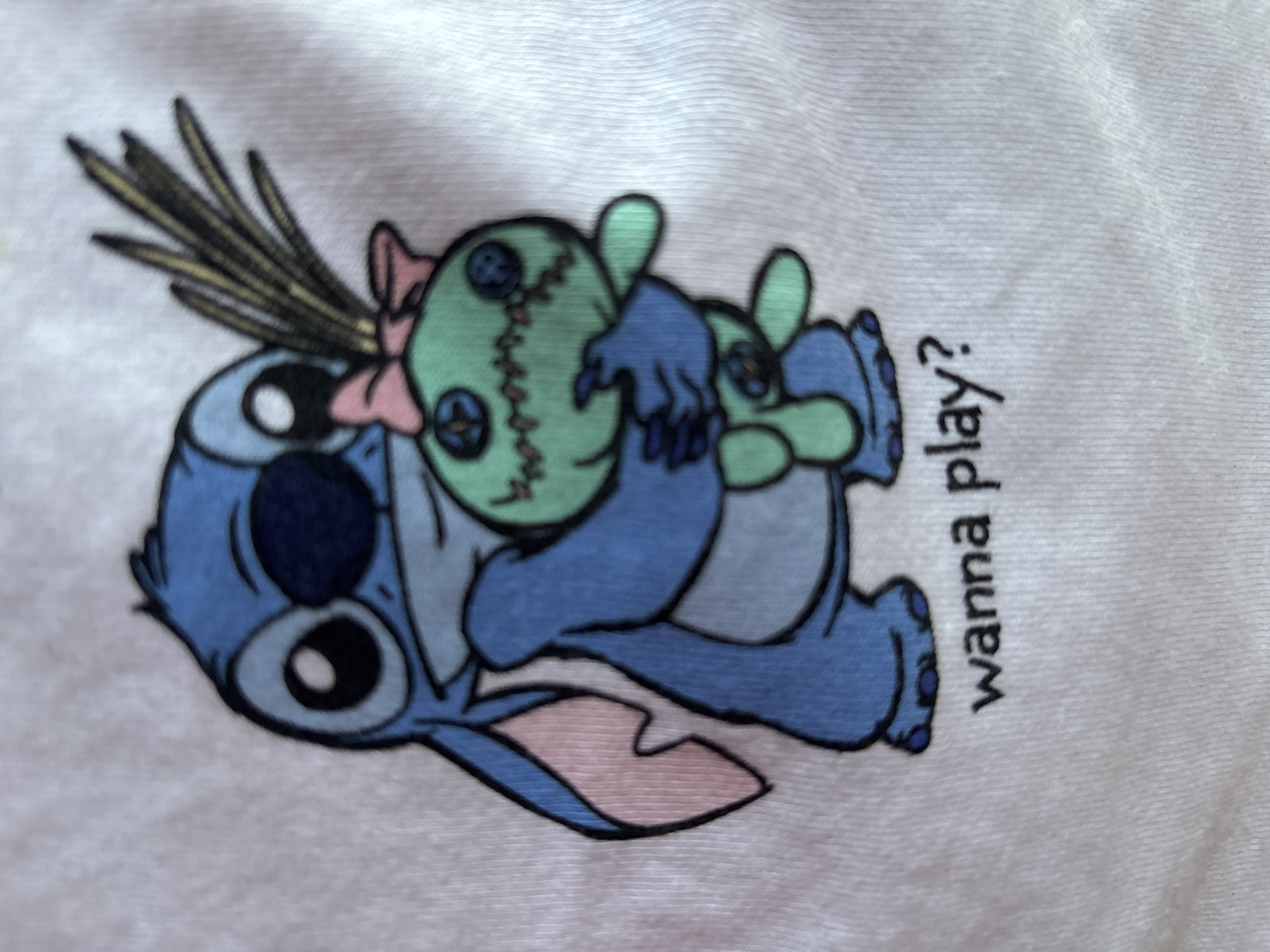 A white t-shirt features a printed image of Stitch, a blue alien character from Disney's Lilo & Stitch, hugging a green voodoo doll. Stitch is depicted with large, black-and-white eyes, blue skin, and pink inner ears. He is holding the voodoo doll close. The voodoo doll is green with black button eyes, stitched X's for a mouth and nose, and a pink bow tied at the top of its head. Several thin, yellow, grass-like strands are sticking out from behind the doll's head. Below the illustration, the text "wanna play?" is printed in black. The t-shirt is laid flat, and the background is plain white with visible fabric texture. The image is focused on the printed design.
