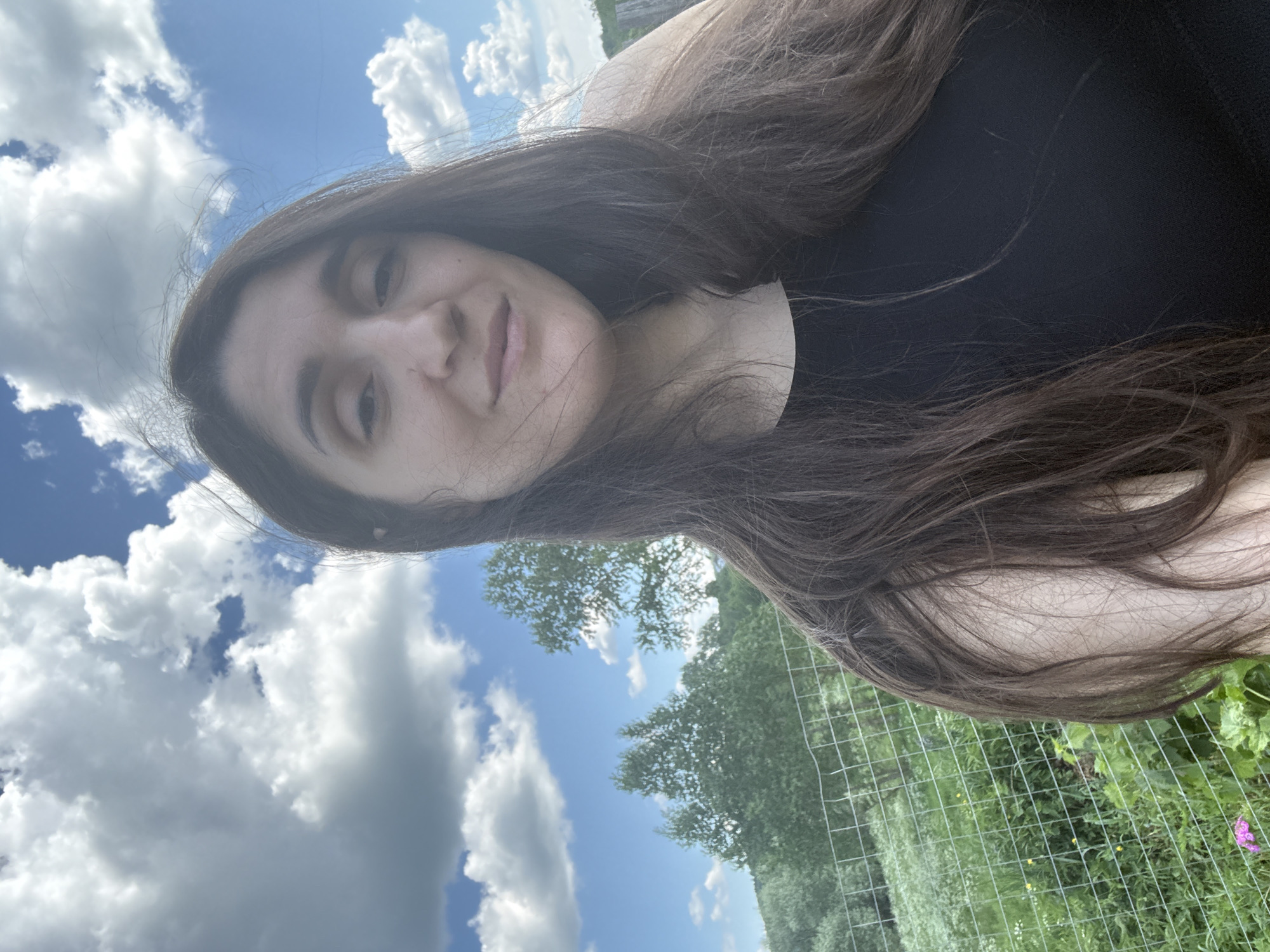 The image shows a woman taking a selfie outdoors. She is in the foreground, her face and upper body visible. She has long, dark brown hair that is partially obscuring her face and shoulder. She is wearing a black tank top. The background consists of a blue sky with white, fluffy clouds, green trees, and a wire fence. The overall impression is of a sunny day in a rural or semi-rural area. The image does not provide any specific details to suggest it was taken in the city of Уральское, Russia.