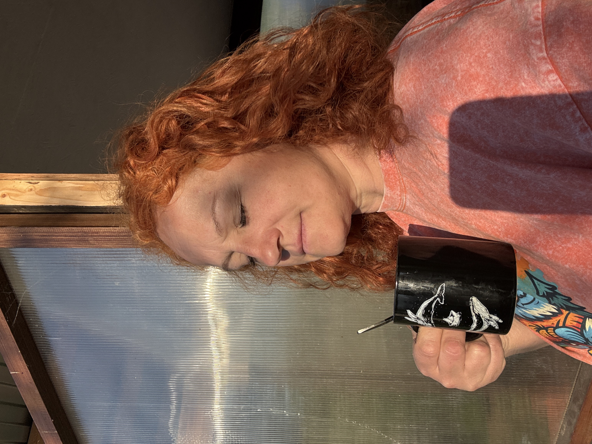 A woman with red curly hair is lying on a translucent corrugated surface with a wooden frame. She is wearing a pink t-shirt and holding a black mug with white whale designs. The scene appears to be indoors, possibly in a greenhouse or sunroom. The image does not contain sufficient information to determine its location in Chaikovsky, Russia.