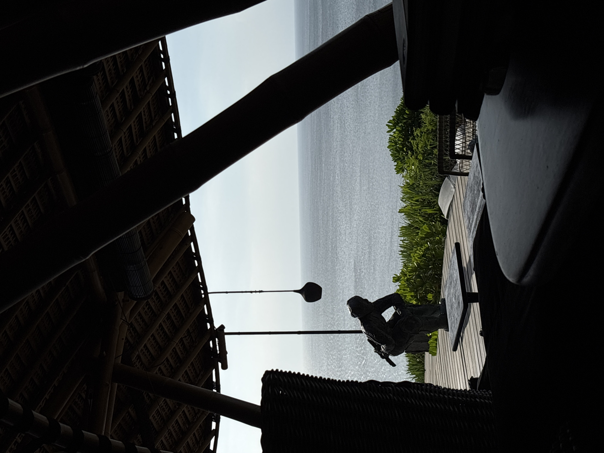 Captured from within a thatched-roof structure in Kuta Selatan, Indonesia, the image reveals a view of the ocean under a bright sky. A wooden deck with green foliage sits to the right, partially obscured by a table. The scene is framed by dark architectural elements of the building, and on the deck is a statue with a long paddle near the roof.