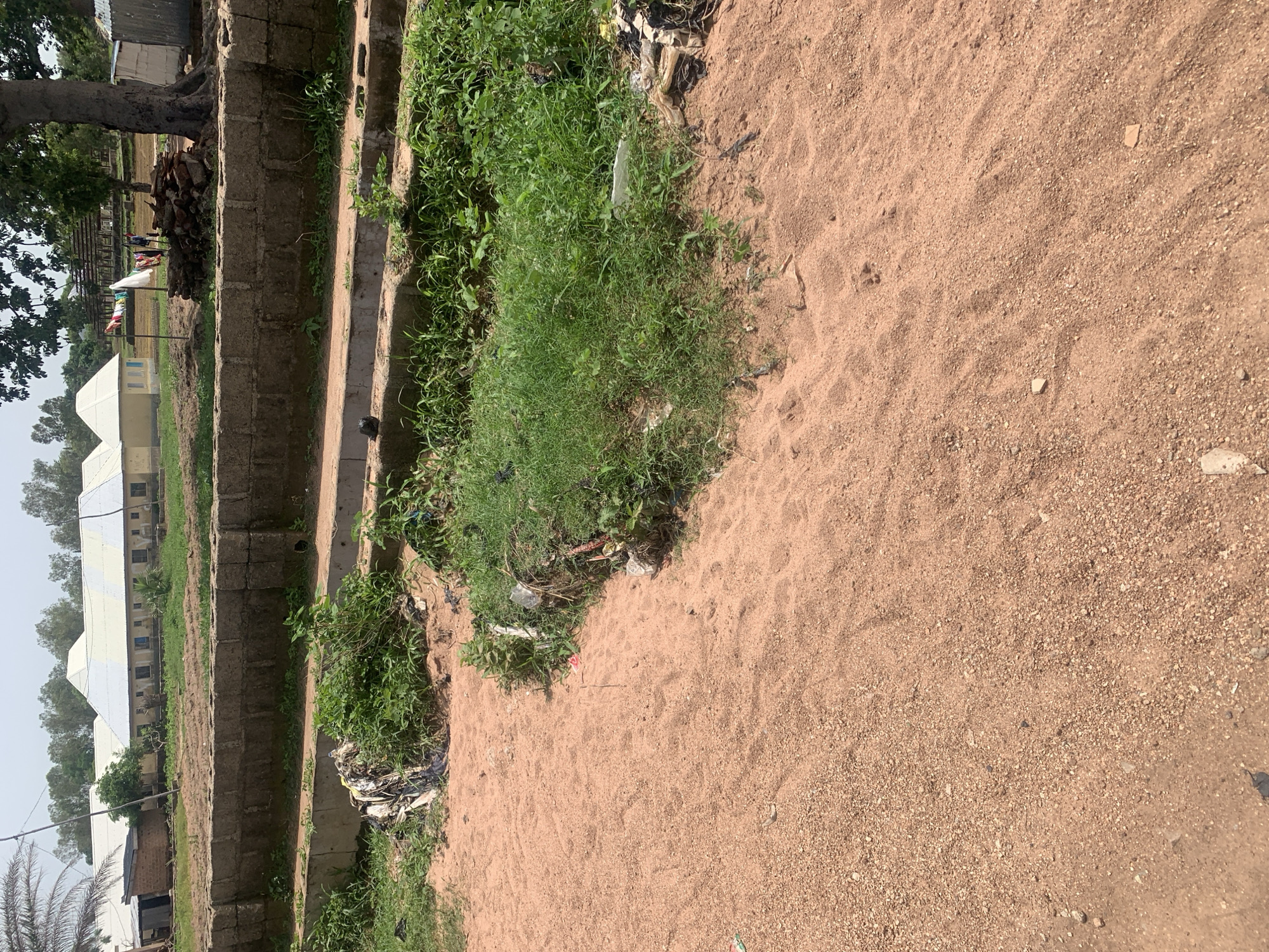 The image depicts an outdoor scene in Jauro Yinu, Nigeria. In the foreground, a sandy area with tire tracks is visible. To the right and slightly behind the sand is a patch of dense green grass. A concrete structure, possibly a drainage ditch or low wall, runs horizontally across the midground, separating the sandy foreground from a brick wall beyond. The brick wall is constructed from rough, rectangular blocks and appears to be a boundary. Behind the brick wall, there is a field with some sparse vegetation. In the background, several buildings with white, sloping roofs are visible. One building has a small annex attached to its left side. Further in the distance, trees are scattered across the landscape, and some laundry is hanging on a clothesline between two poles in front of a shaded structure. The overall impression is of a rural or semi-urban environment under a bright, overcast sky. There are no people visible in the image.