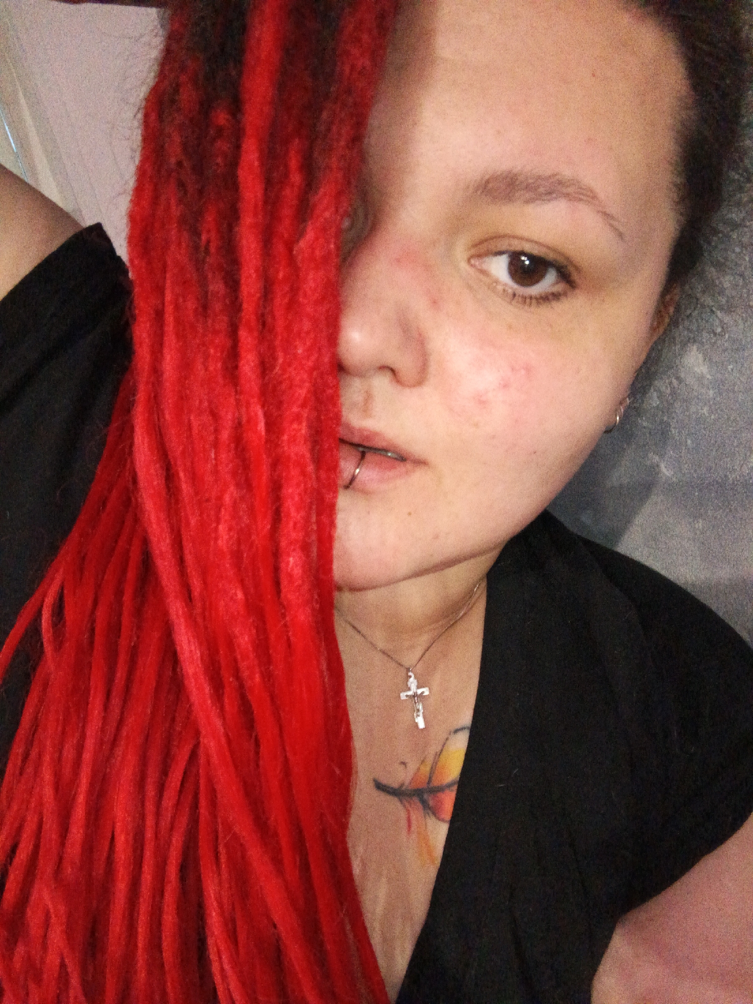 The image depicts a close-up, chest-up portrait of a single female individual in Chaikovsky, Russia. The subject has vibrant red dreadlocks, which obscure the left side of her face. Her visible right eye is open, looking directly at the camera. Her skin shows some minor redness. A silver-colored labret ring is present in her lower lip, and a small silver hoop earring is in her right ear. She wears a silver-colored necklace with a crucifix pendant resting on her upper chest. A colorful tattoo, appearing to be a feather design with hues of yellow, orange, and red, is visible on her upper chest beneath the necklace. She is dressed in a dark, possibly black, garment. The background consists of a textured, light grey surface, likely an interior wall. The subject appears to be taking a self-portrait.