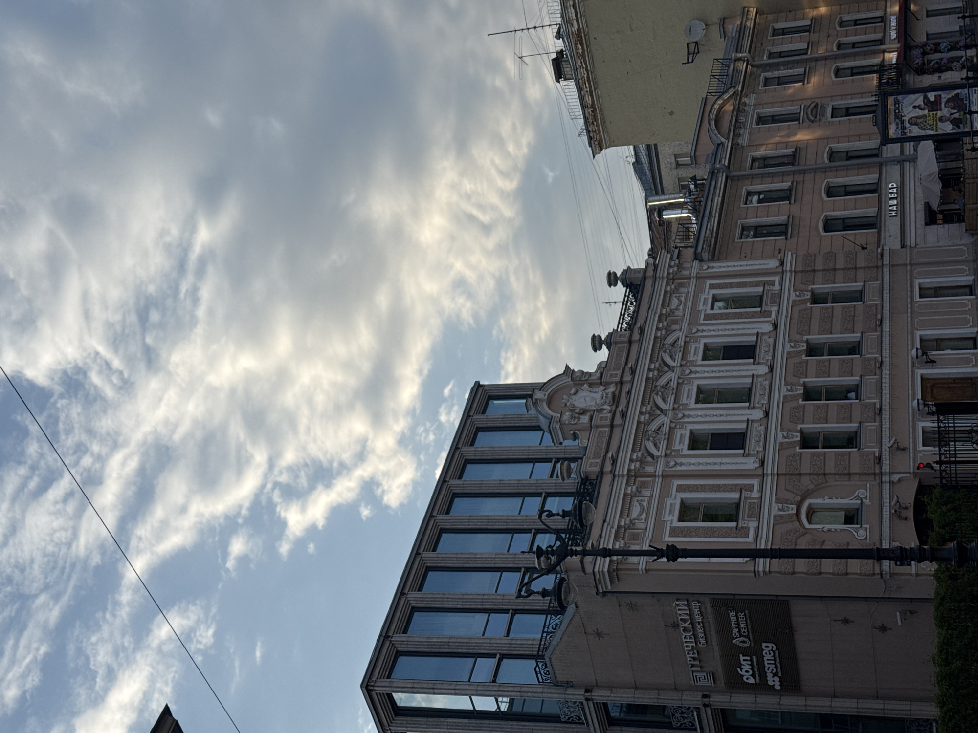 Here is a description of the image:
The image captures an upward angle shot of a building facade against a cloudy sky. The building has a classical architectural style with ornate details, multiple windows, and signs on the lower part. Signs on the lower part of the building facade read "ГРЕЧЕСКИЙ Бизнес Центр", "Sapphire Center", "ОБИТ", and "smeg". A streetlamp is also visible in the foreground. The sky is filled with white clouds, suggesting either early morning or late afternoon. The building appears to be in an urban environment, likely located in Saint Petersburg, Russia due to the Cyrillic script on the signs. There are no people visible in the image.
