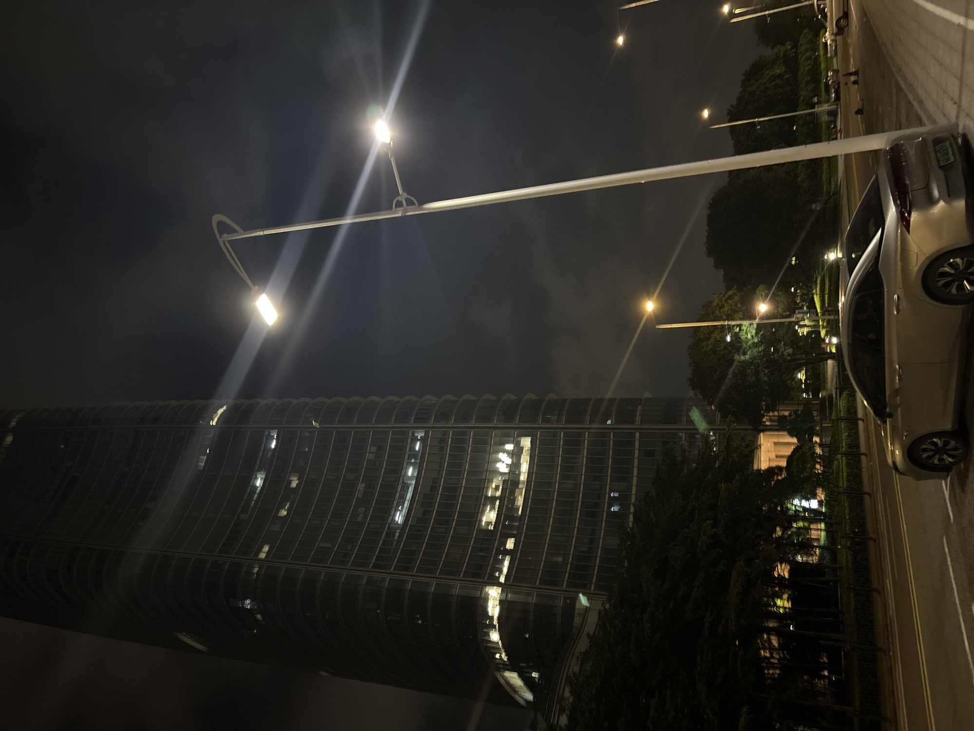 This media file captures a nighttime scene in Haizhu District, China. The dominant subjects are a modern, curved skyscraper with illuminated windows and a street lined with trees and streetlights. A silver car is parked on the right side of the road. The activity is a quiet urban evening, with the city lights reflecting on the wet pavement.
Notable details include the dark, cloudy sky, suggesting a humid or overcast night. The streetlights cast bright beams, creating lens flares in the image and illuminating the scene. The skyscraper's architecture is a prominent feature, its curved facade and grid of windows creating a sense of scale and grandeur. The trees are lush and dark, contrasting with the man-made structures. The presence of multiple streetlights and the car indicate a public area, likely a street or roadside park adjacent to the building. No people are clearly visible, contributing to a sense of peaceful solitude. The overall emotion conveyed is one of serene urban nightlife. No visible text is present in the image.